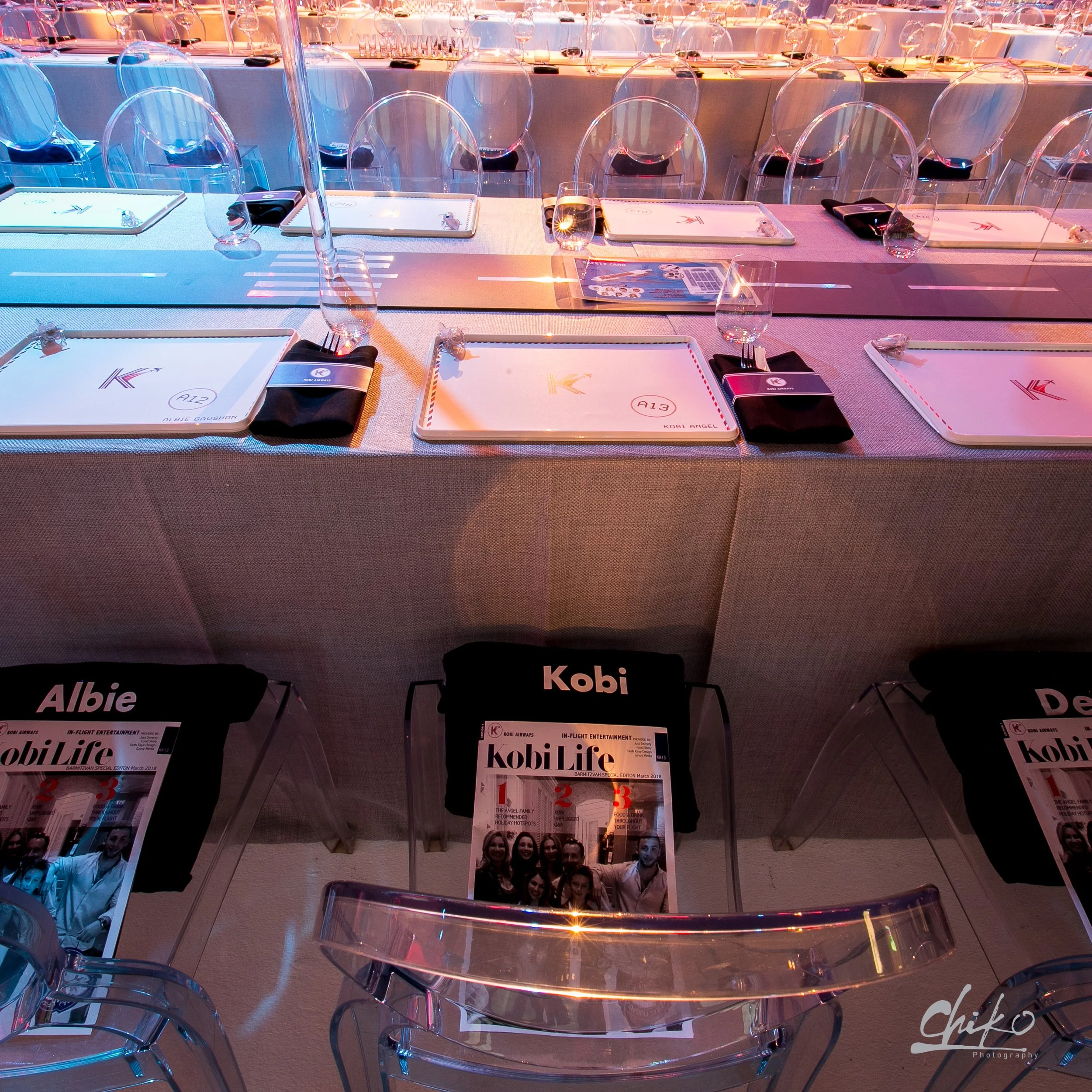 A long banquet table set for a formal event, with transparent chairs, placemats, glasses, and utensils arranged neatly. There are magazines or programs on the table with the titles 'Kobi Life' and magazine covers featuring groups of smiling women.