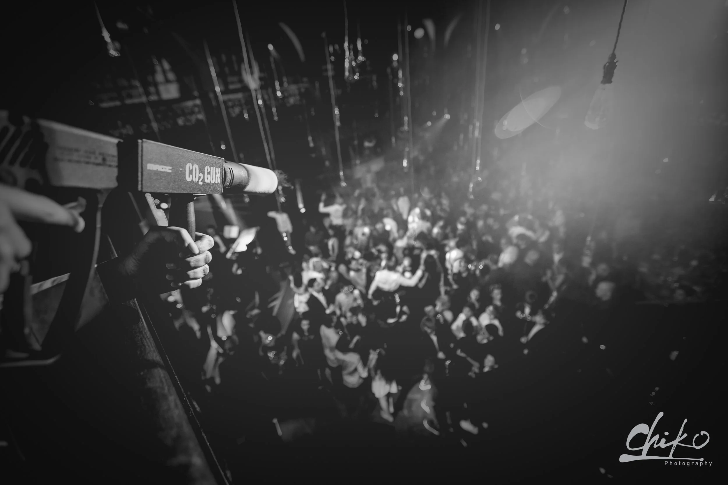 A crowded scene with a person holding a CO2 gun in the foreground, shooting into a large crowd at a nightclub or concert. The scene is in black and white, with spotlights and blurred figures of people.