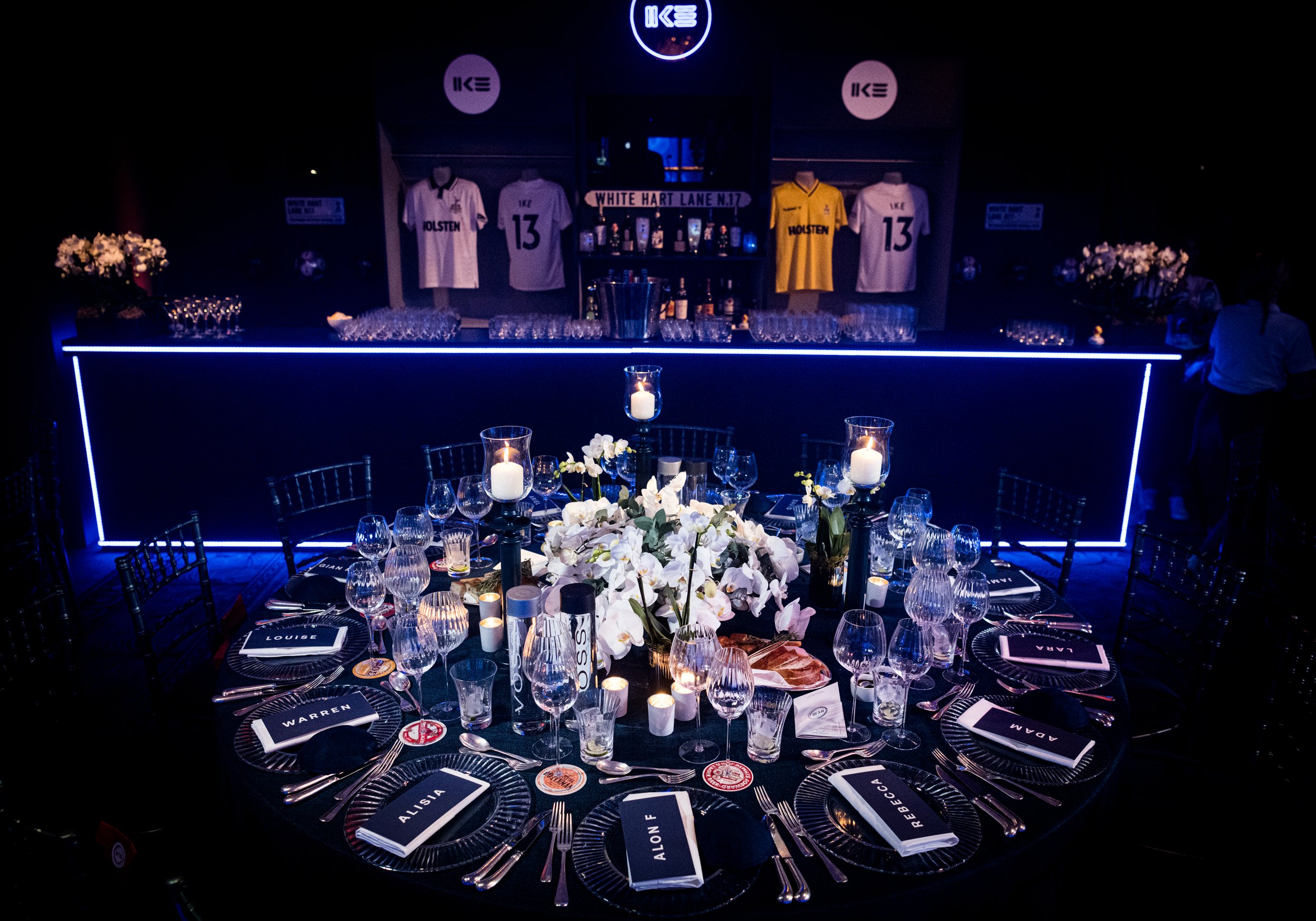 A dimly lit banquet table decorated for an event, with a centerpiece of white flowers and candles, arranged with glasses, utensils, and personalized name cards for each guest. The background features a bar with sports jerseys on display and neon sign