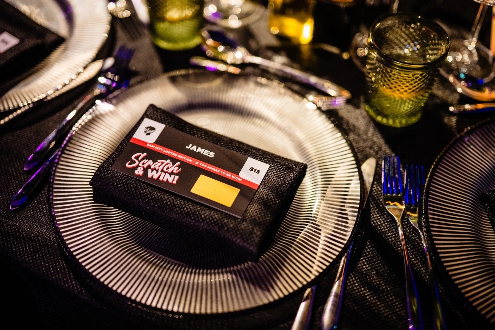 A table setting with a black and white striped plate, silverware, a green glass, and a menu with a scratch card on top, all arranged on a black textured tablecloth.
