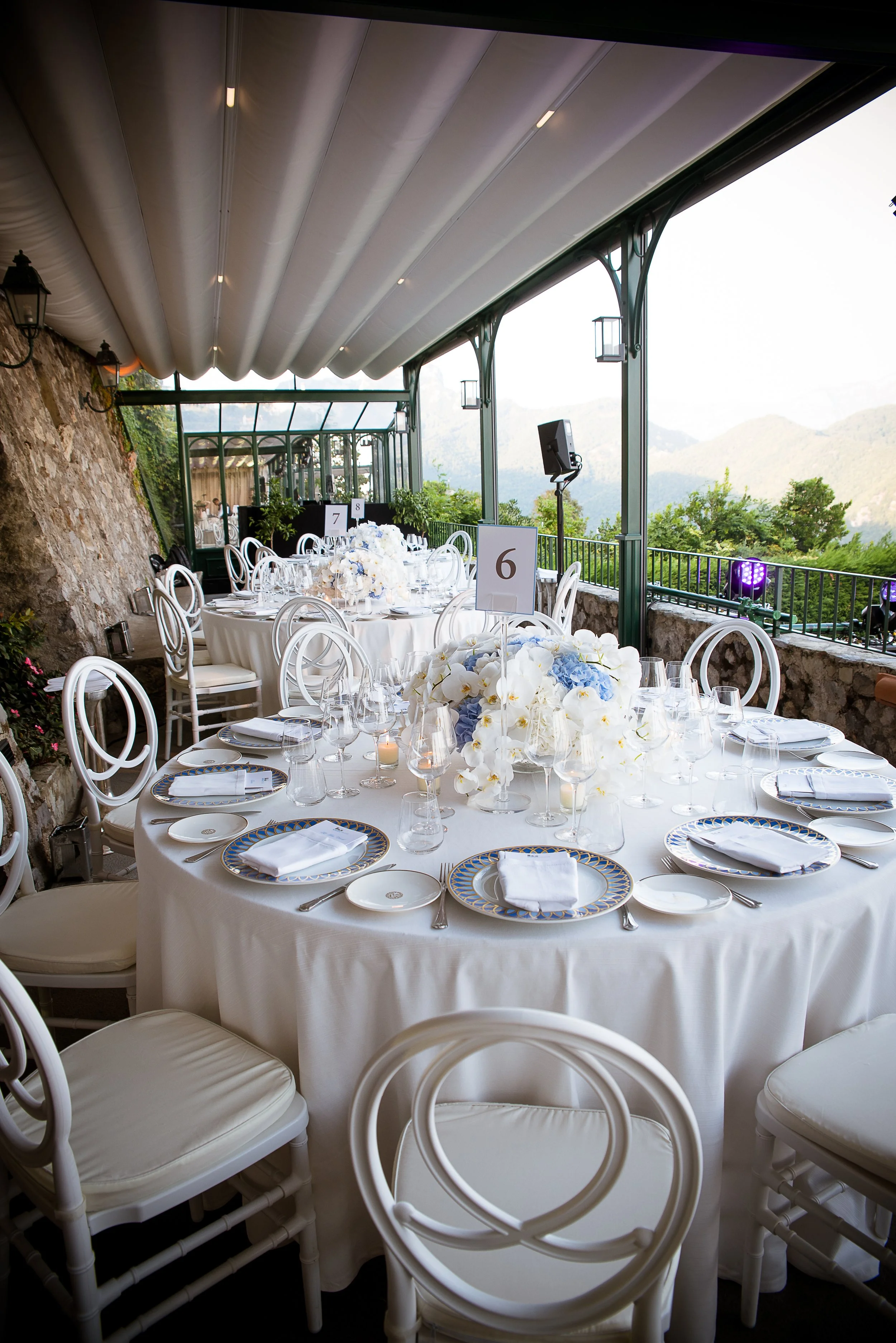Elegant outdoor banquet setup with white tablecloths, floral centerpieces, and white chairs on a terrace overlooking mountains, with a canopy overhead and string lights