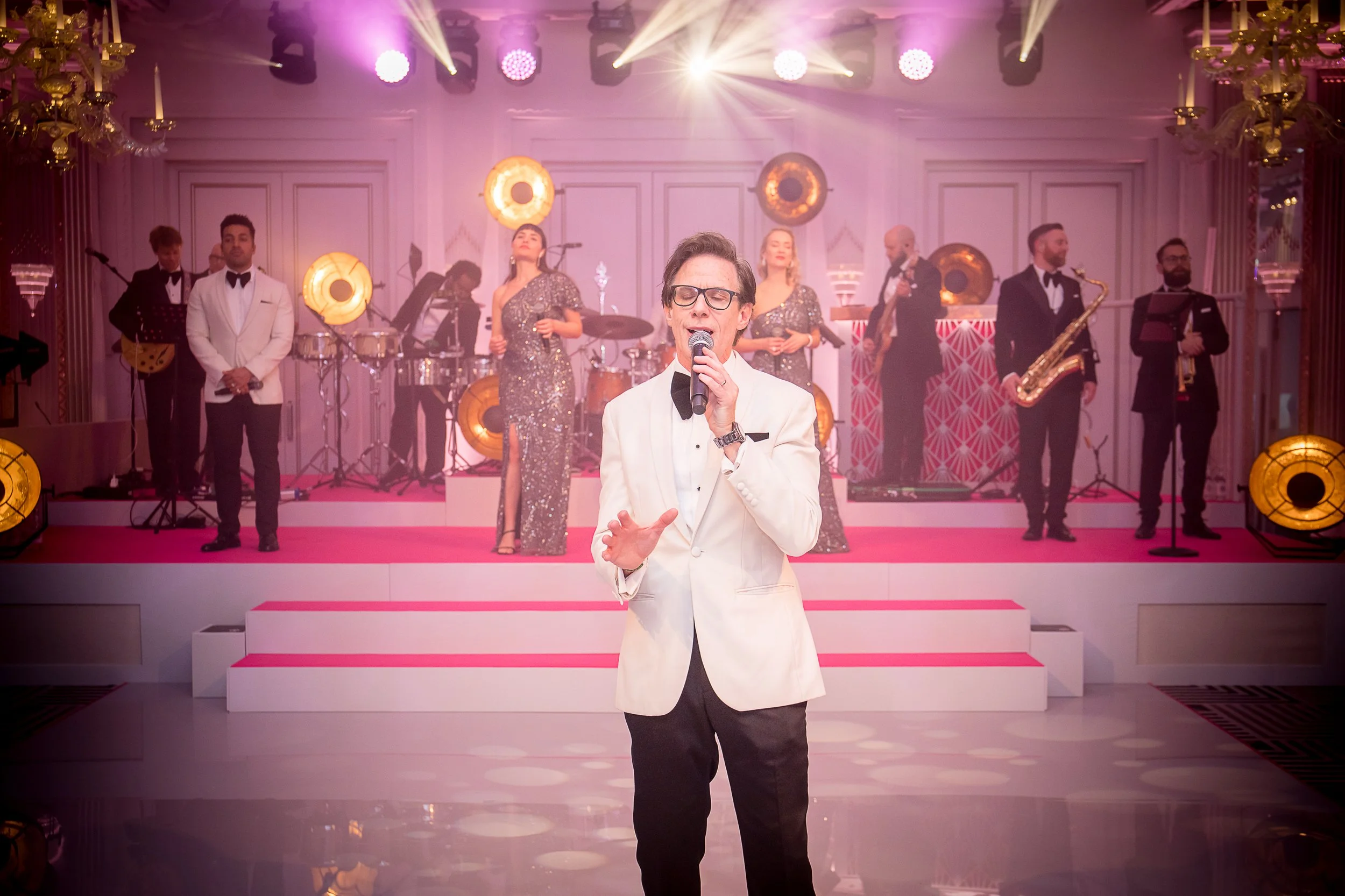 A man in a white tuxedo jacket and black pants singing into a microphone in the foreground, with a band and backup singers dressed in formal attire performing on a stage decorated with pink and gold lighting in the background.