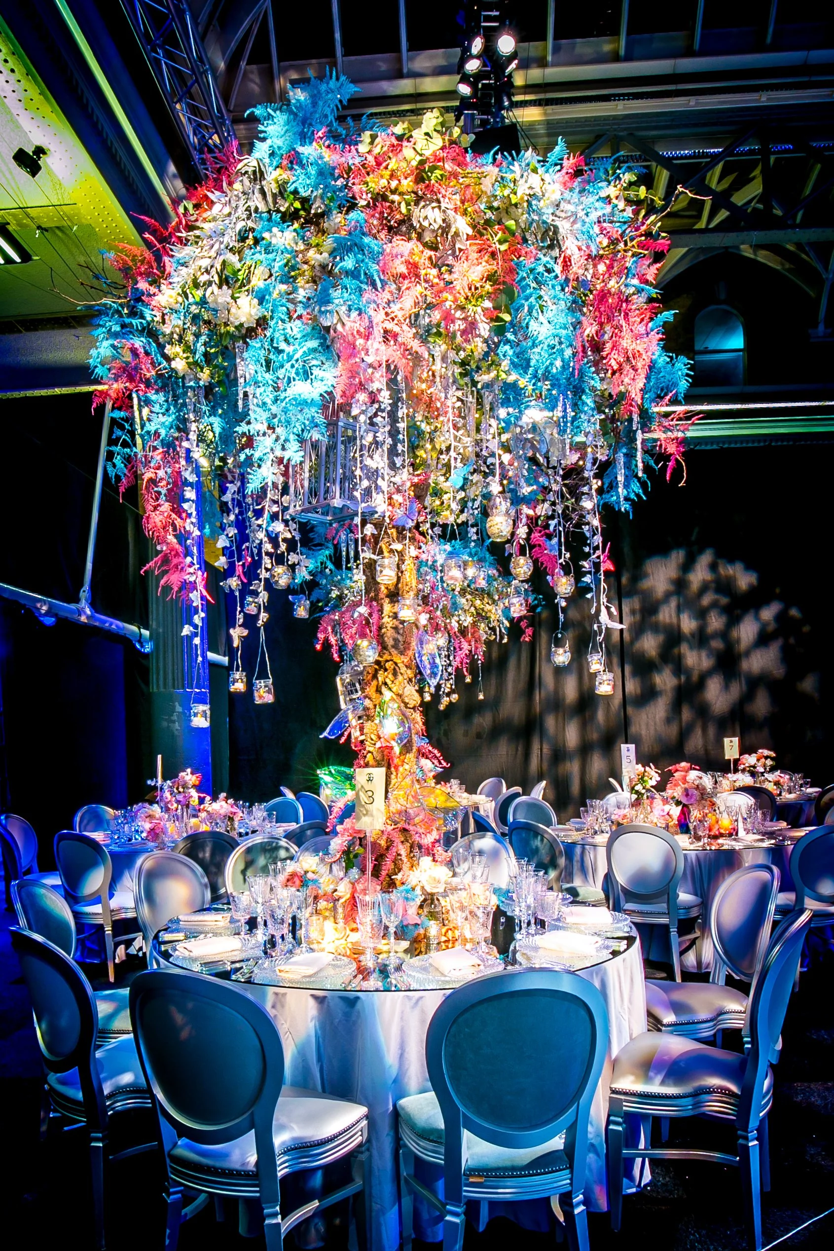 Elegant banquet hall decorated with a tall floral centerpiece with colorful flowers and hanging glass jars, surrounded by round tables with white tablecloths, fine china, and chairs, illuminated by vibrant blue lighting. ICE PALACE BAT MITZVAH at Old