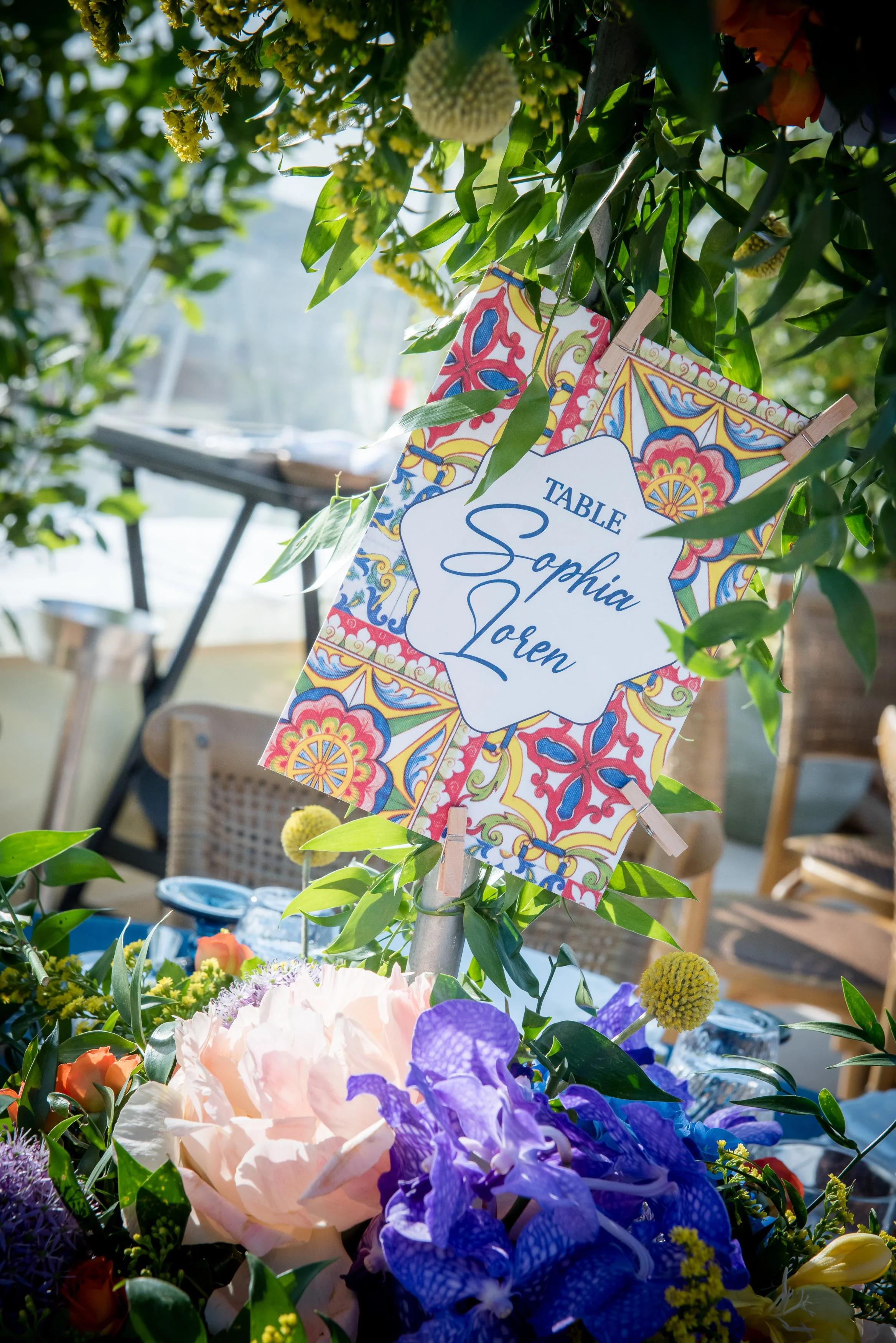 A decorative sign with the name 'Sofia Loren' hanging among colorful flowers and green leaves at a table set up outdoors on a sunny day.