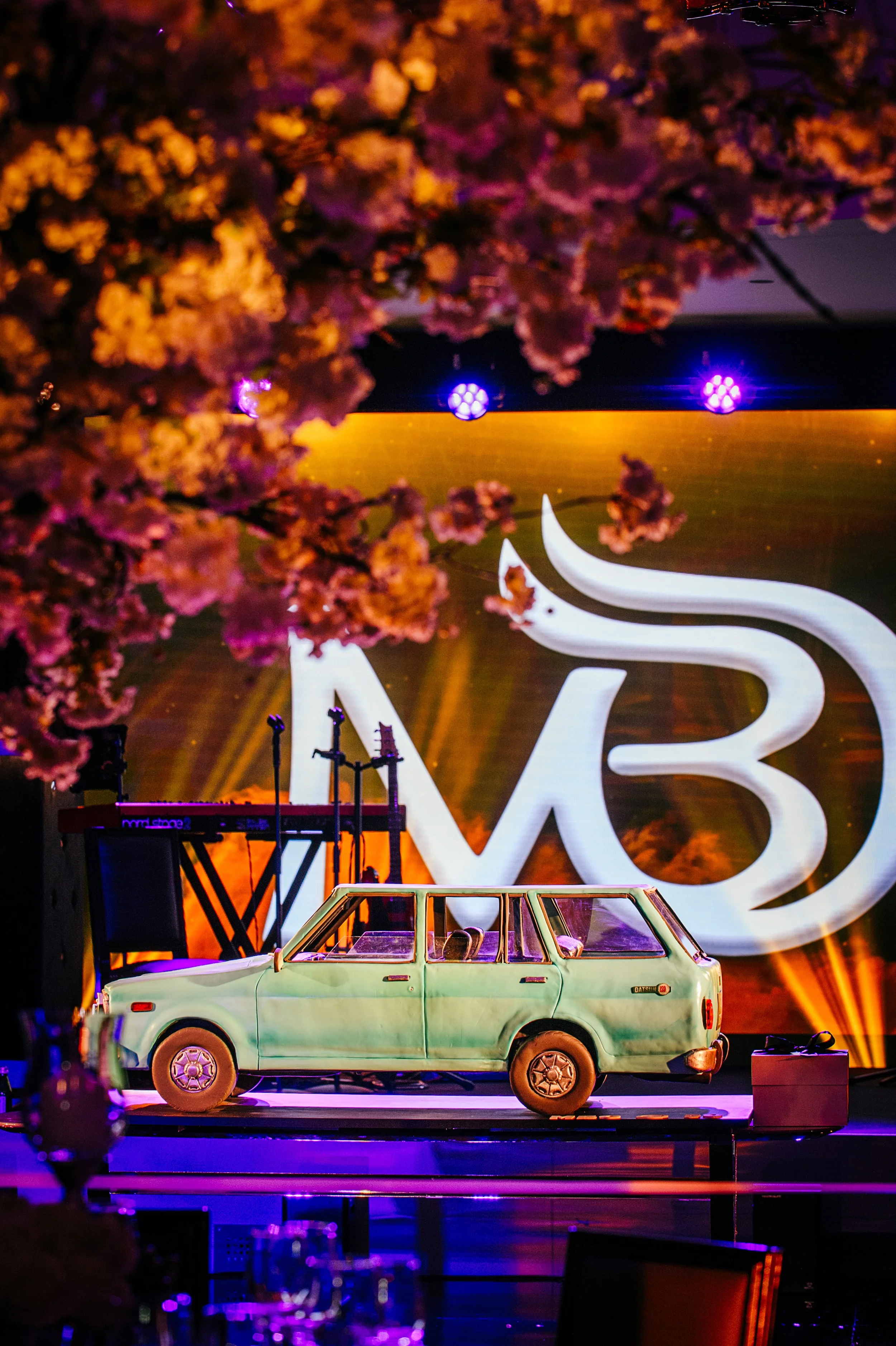 A vintage light green car displayed on stage with pink cherry blossom trees in the foreground and purple stage lights in the background.
