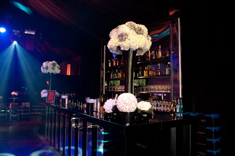 Bar with floral decorations, tall floral arrangements in vases, bottles of liquor on shelves, glassware, and colorful lighting.