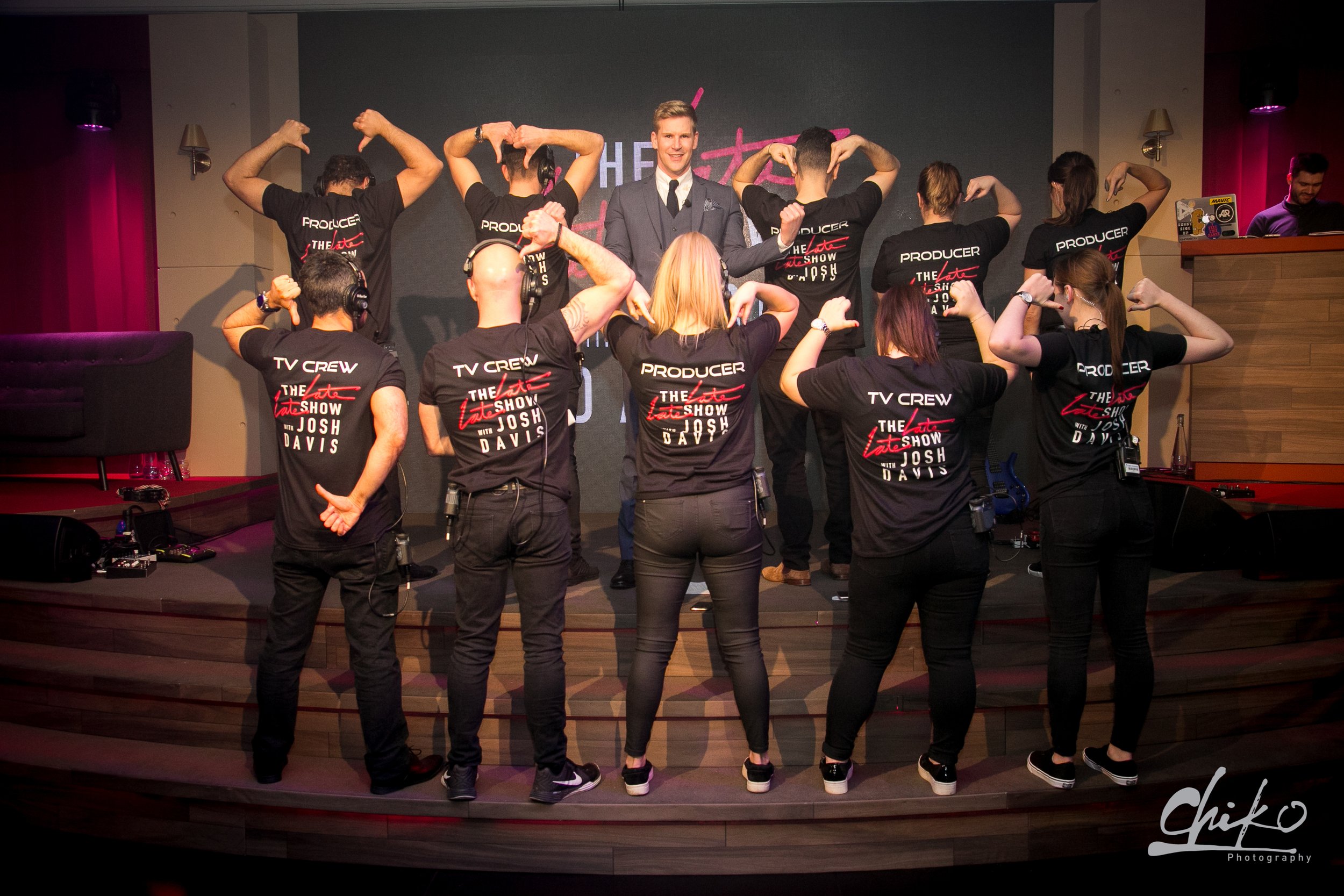 Group of people on a stage taking a selfie, with a man in a gray suit at the center and others wearing black T-shirts with writing on the back, some making muscle poses and others pointing to their heads.