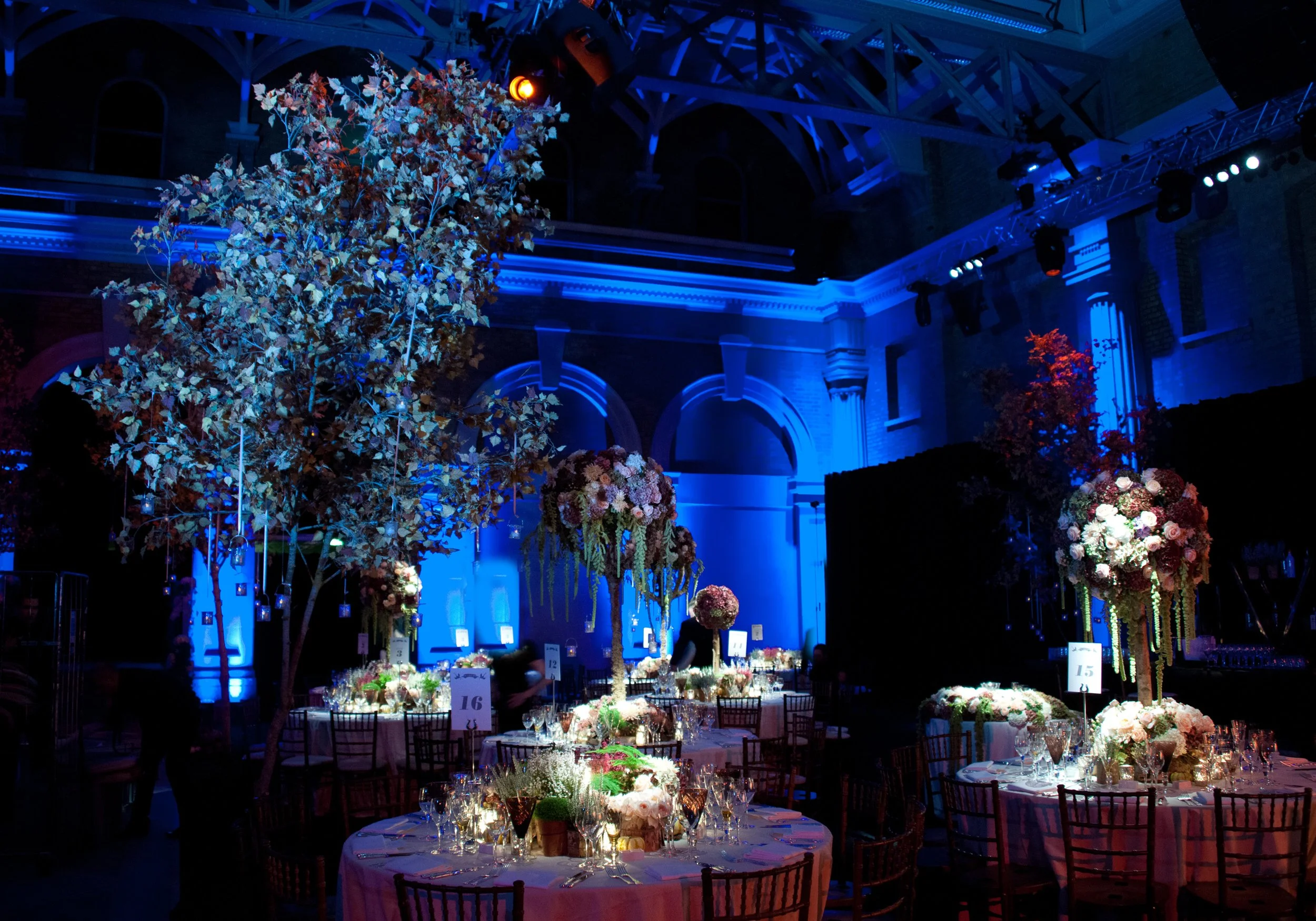 Indoor event space with decorated tables, floral arrangements, and blue lighting, likely set up for a banquet or wedding reception.