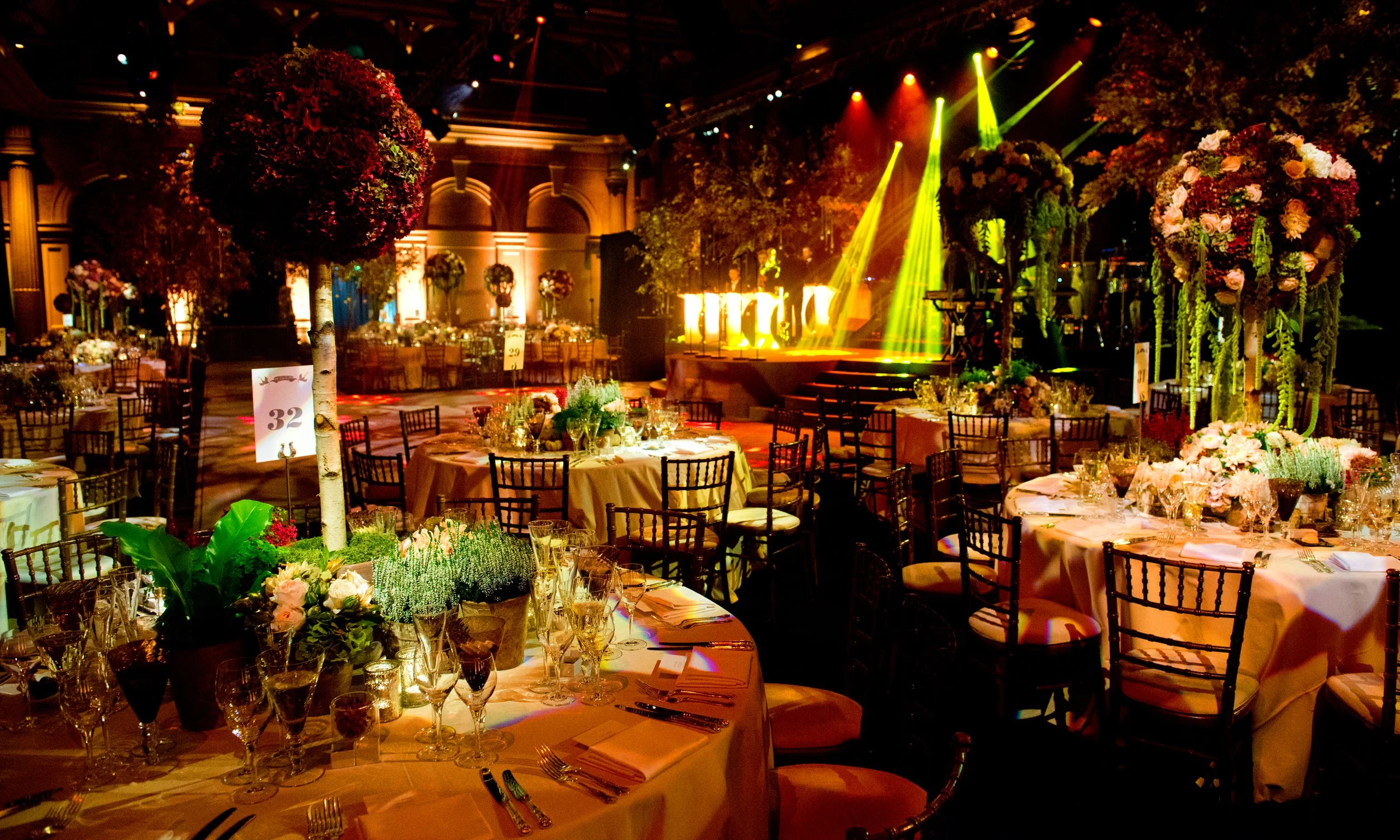 An elegant banquet hall decorated with tall floral centerpieces, round tables set with glasses and cutlery, and colorful stage lighting in yellow, red, and green illuminating the room.