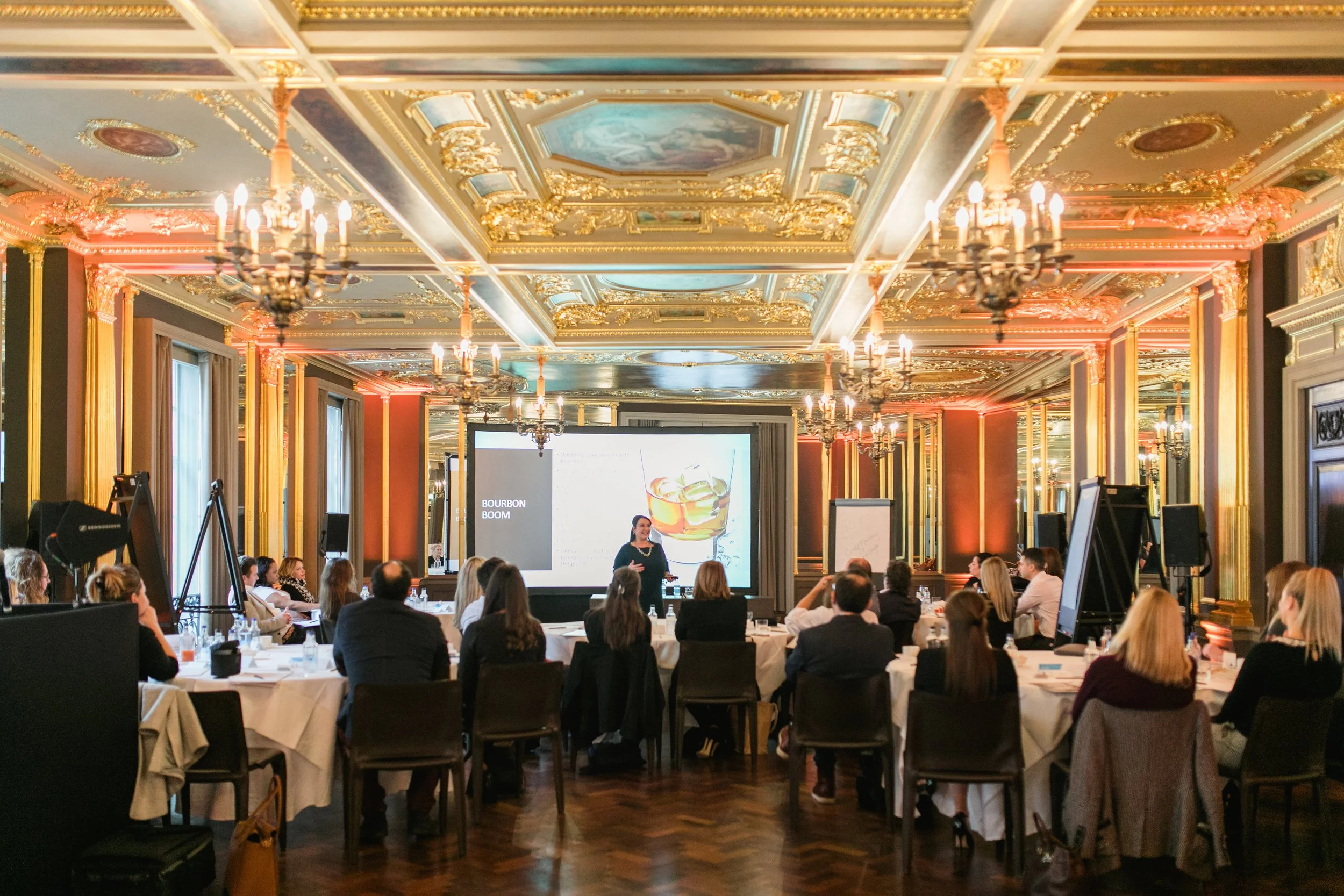 A conference room with ornate gold and pastel decor, chandeliers, and a large screen displaying a presentation on bourbon.