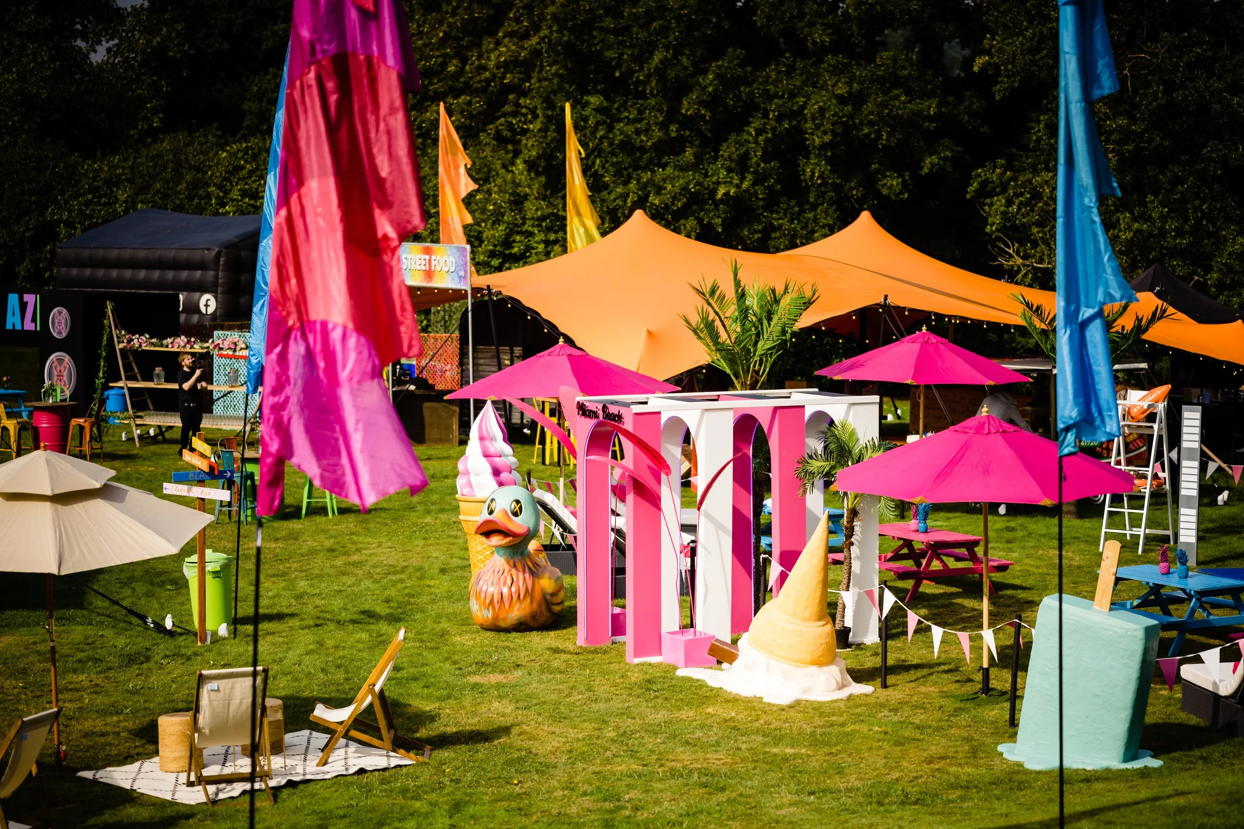 Colorful outdoor festival scene with pink and blue umbrellas, orange canopy tents, decorative palm trees, inflatable ice cream cone, and a sign that reads "Street Food" set on a green grassy area with trees in the background Wrotham Park