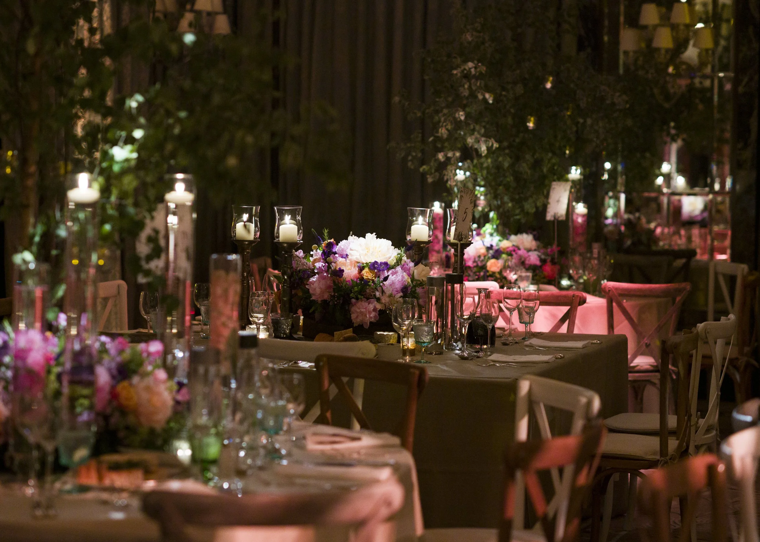 Elegant dining setup with floral centerpieces, candles in glass holders, and wine glasses on tables at an event, illuminated by soft candlelight.