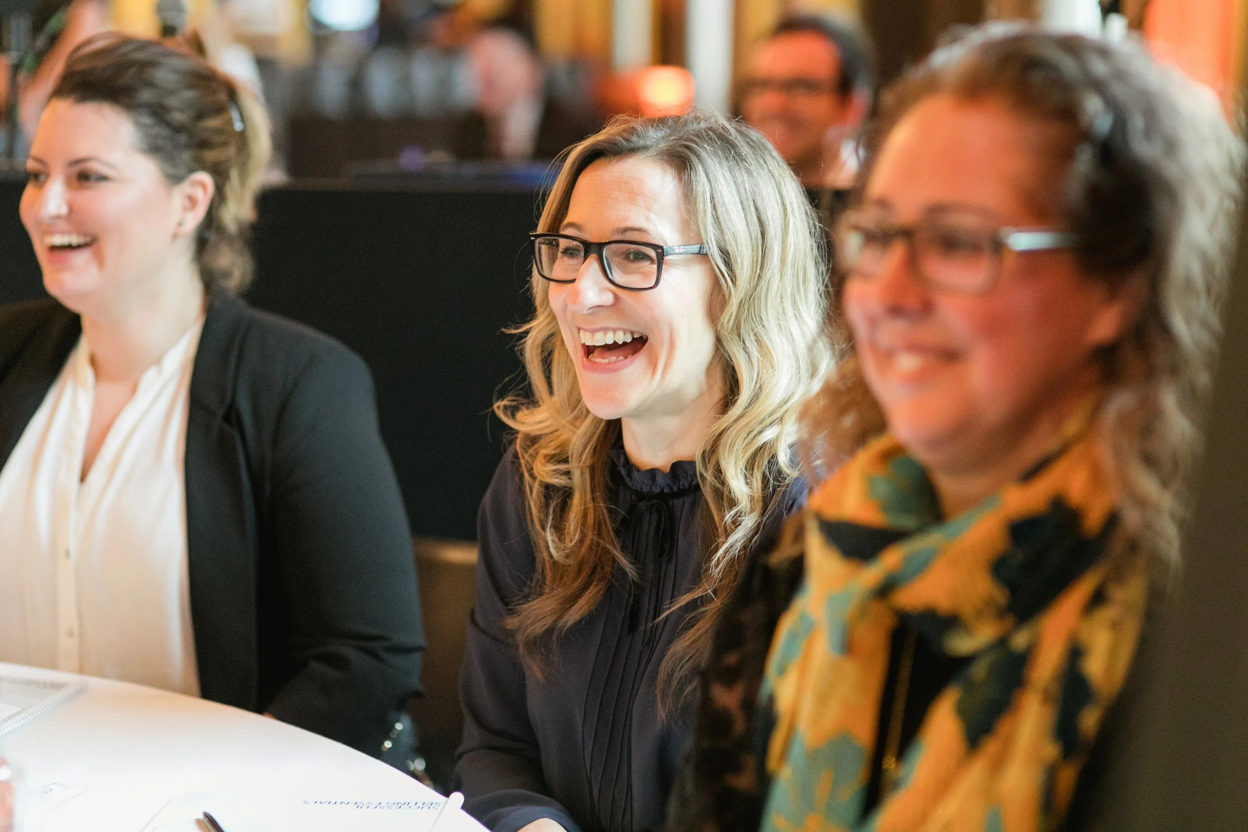 Three women sitting at a table in a social setting, smiling and laughing, with a blurred background of other people and warm lighting.
