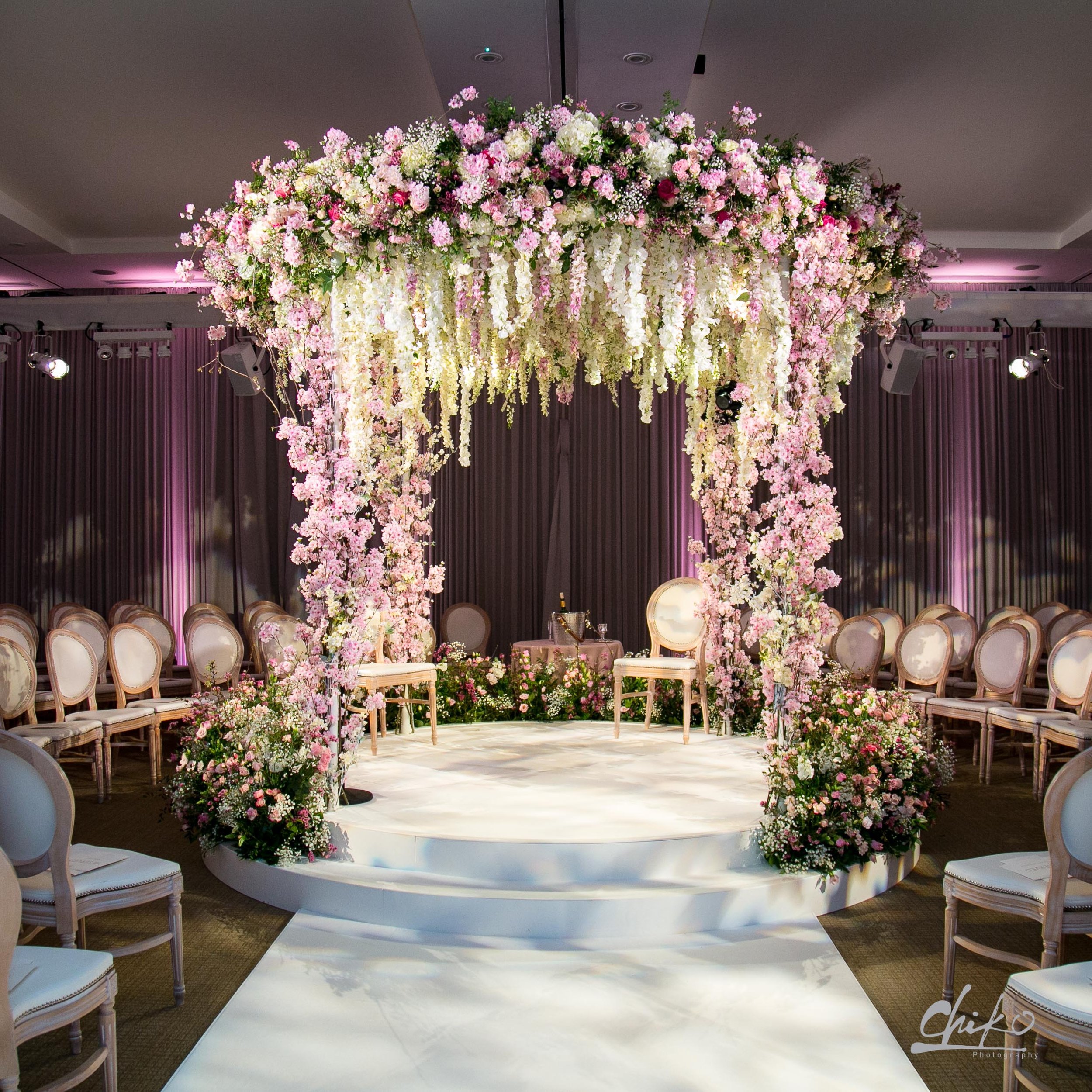 A floral wedding arch decorated with pink and white flowers inside a decorated hall with chairs arranged on either side.