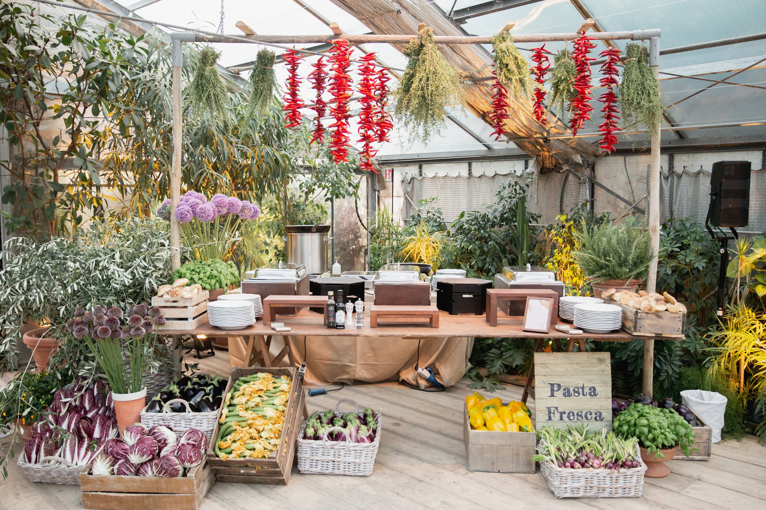 A greenhouse and outdoor market setup with hanging red chili peppers, bunches of herbs, and various fresh vegetables displayed in baskets and crates, with a sign reading 'Pasta Fresca'. Venue is in Tuscany at Serre Torrigiani Bistrot