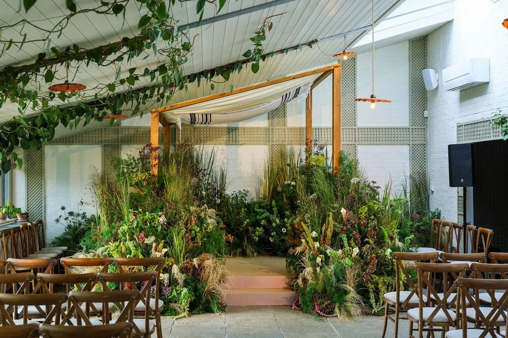 Indoor venue decorated with lush green plants and flowers with wooden chairs arranged on both sides of a small stage, featuring a canopy and string lights.