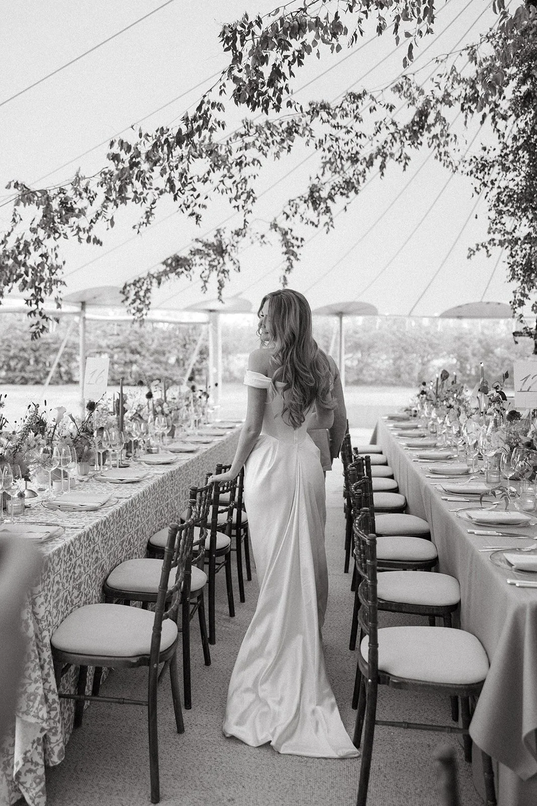 A woman in a wedding dress standing between two long dining tables under a tent with hanging leaves and string lights.