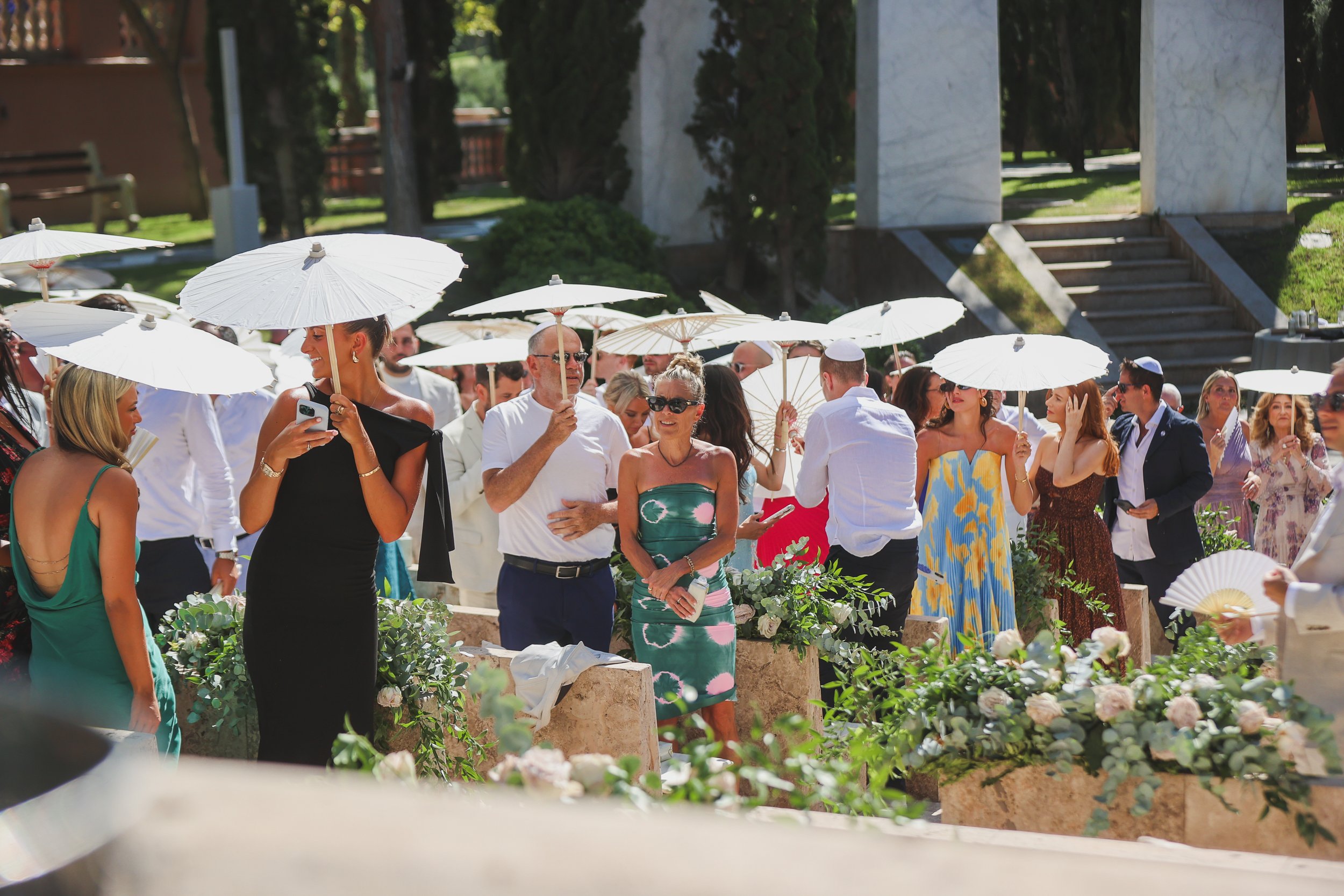 A crowd of people dressed in formal attire participating in an outdoor wedding or celebration in a garden, shaded by white parasols.
