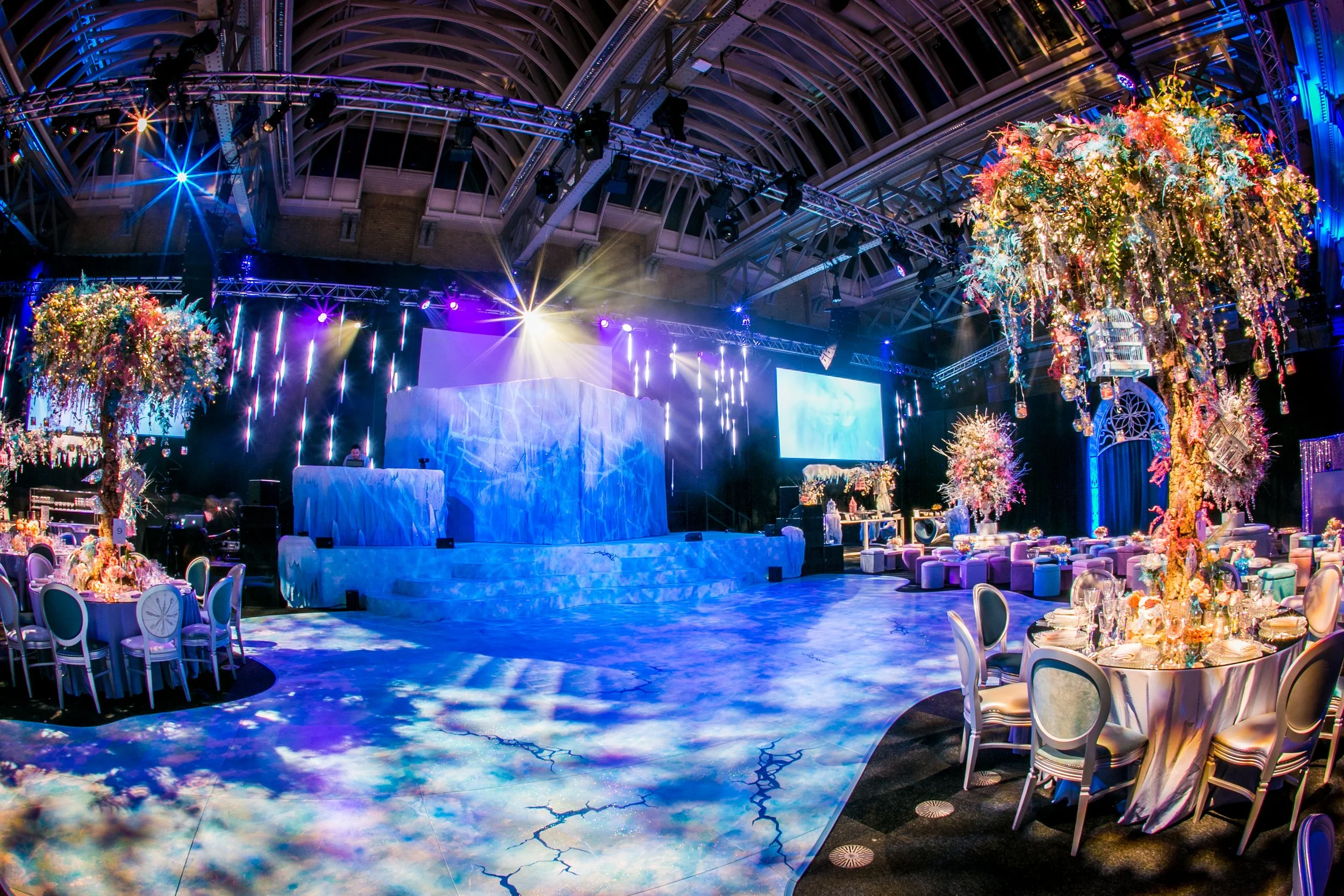 A large event space decorated with colorful floral arrangements, illuminated with vibrant stage lighting and a spacious dance floor, set up for a wedding or celebration. ICE PALACE BAT MITZVAH at Old Billingsgate 