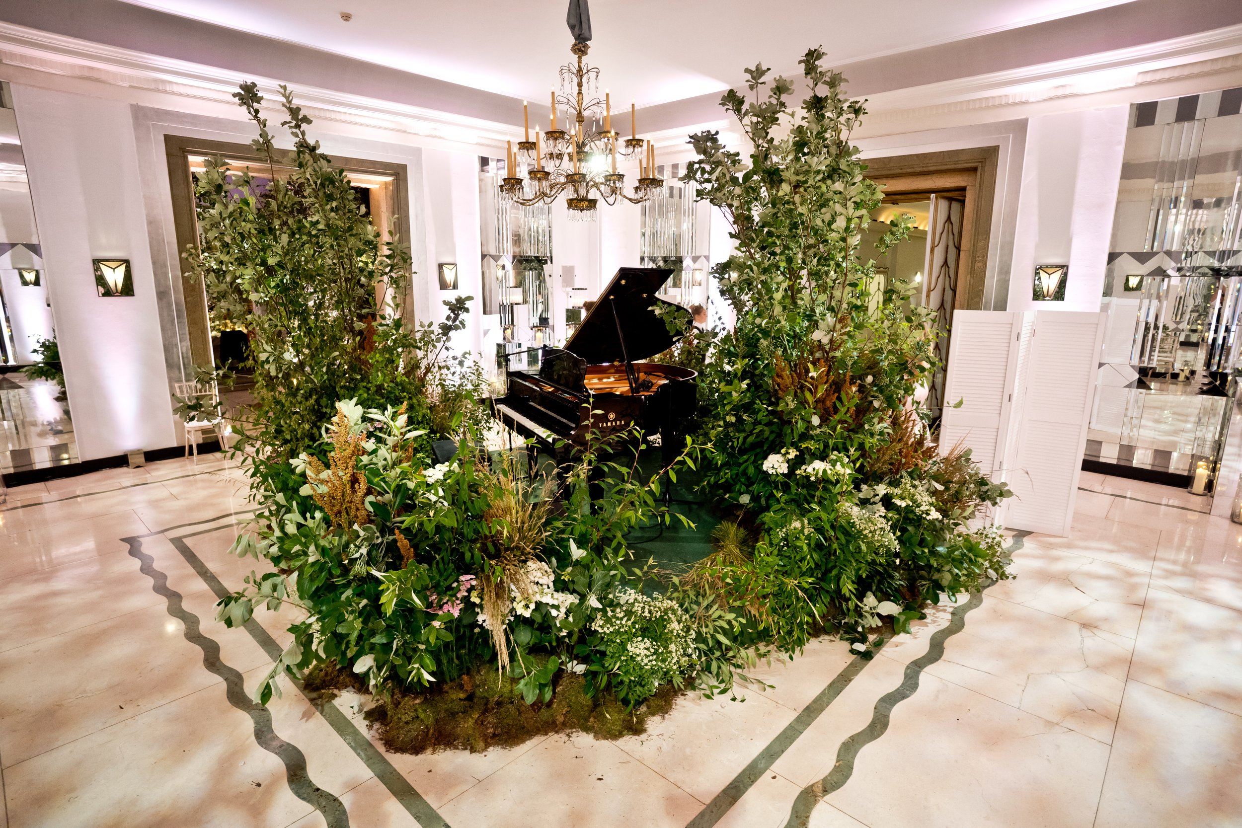 A grand indoor setting with a lush green floral arrangement surrounding a grand piano, a chandelier hanging from the ceiling, and elegant interior decor.