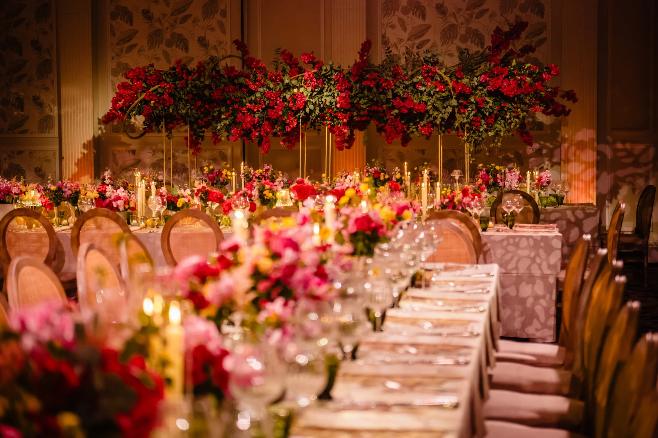 An elegant banquet table decorated with pink, red, and yellow flowers, tall candles, and glassware, set in a room with floral wallpaper and soft lighting, suitable for a celebration or wedding banquet.