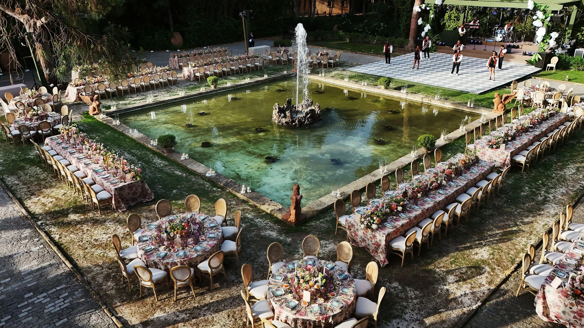 An outdoor banquet setup with round tables covered in floral tablecloths and floral centerpieces, arranged around a rectangular fountain with a central water fountain, set in a garden area with lush greenery, trees, and a stage with musicians playing instruments in the background.