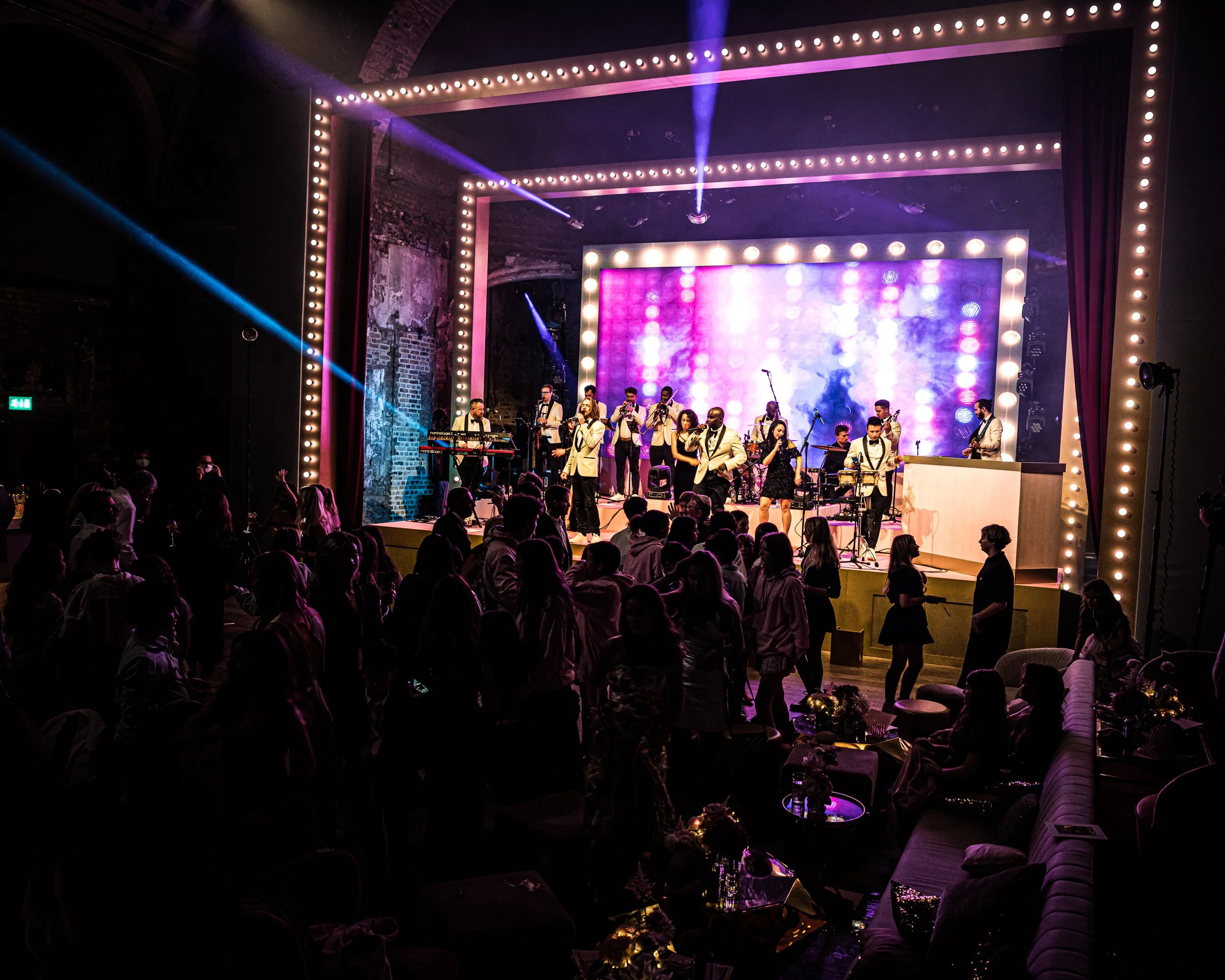 An indoor concert stage with a full band performing for an audience. The stage is decorated with bright lightbulbs around the border and purple and blue lighting effects.BAT MITZVAH AT BATTERSEA ARTS CENTRE