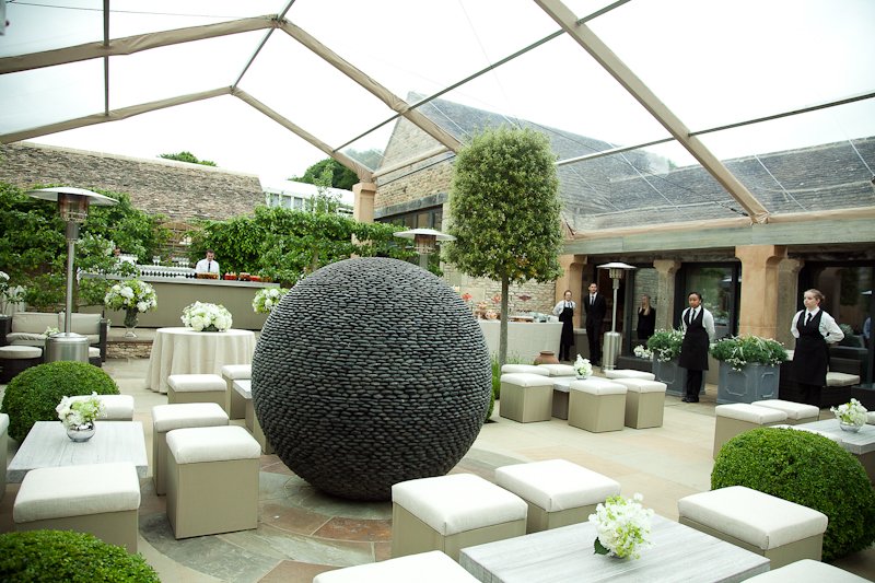 Outdoor patio with lounge chairs, small tables with flower arrangements, a large black spherical sculpture, well-manicured bushes, and waitstaff in black uniforms