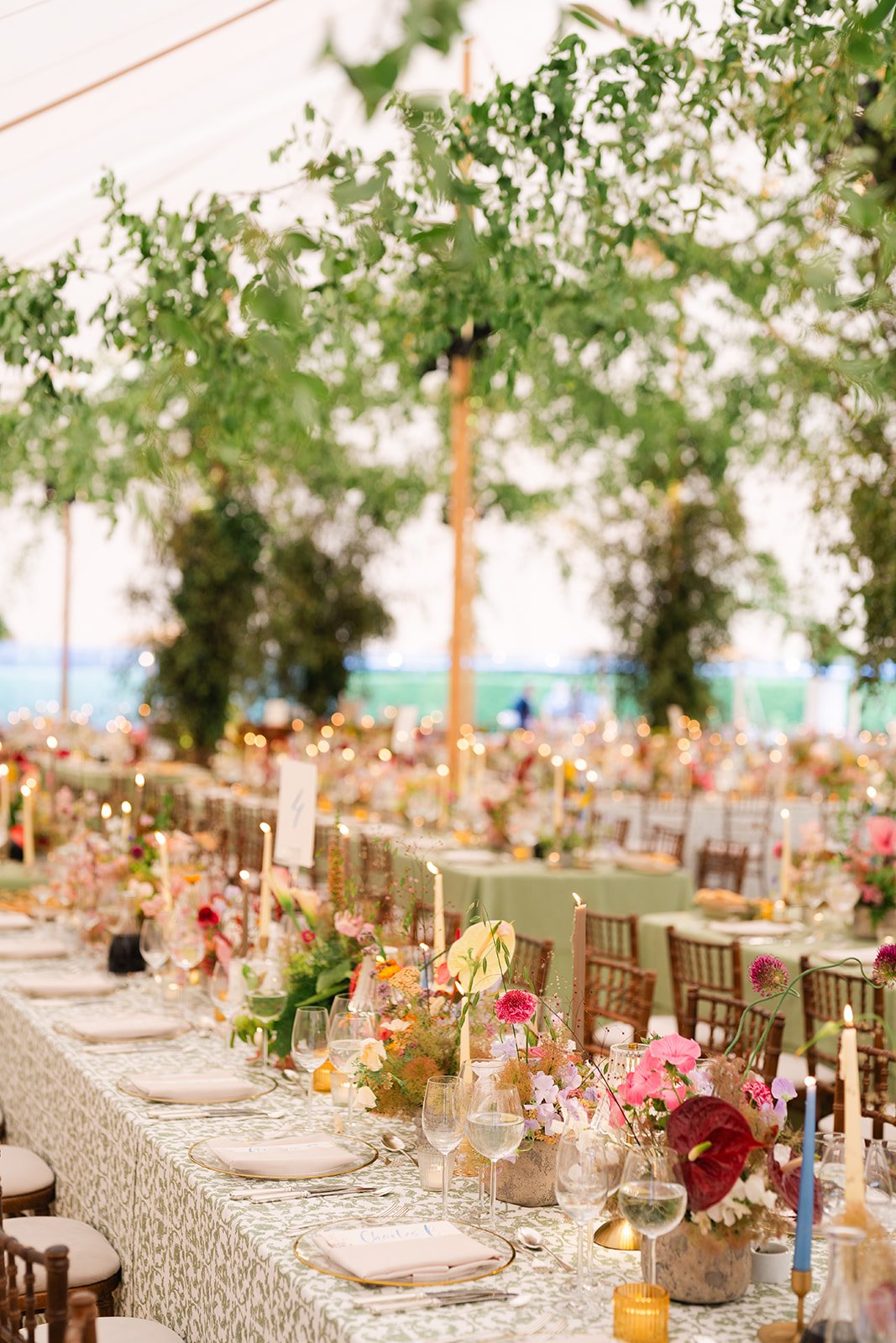 Elegant event venue with a long rectangular table decorated with floral centerpieces, candles, and tableware, under a white canopy with hanging green foliage.