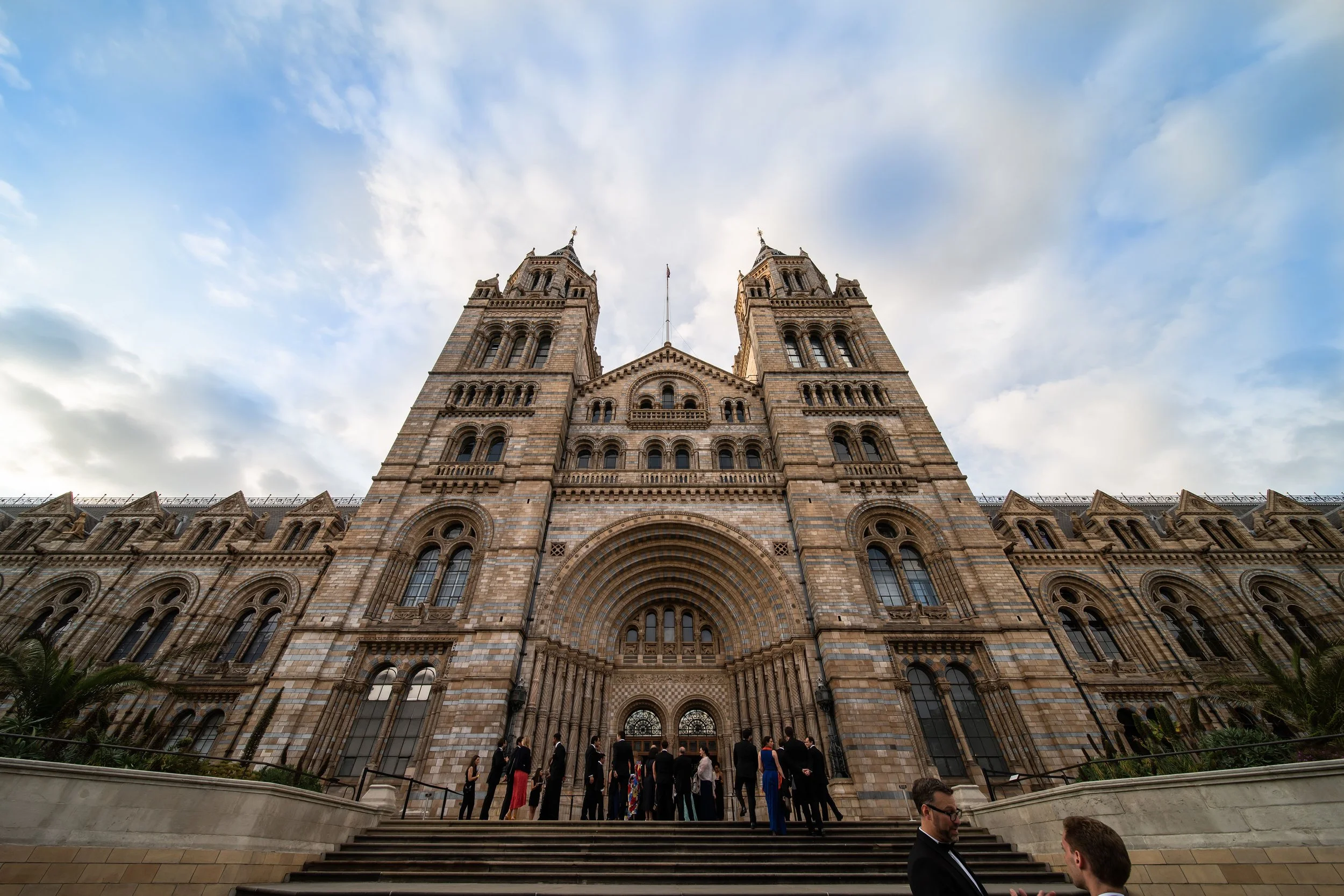 The Natural History Museum with corporate guests in formal attire gathering outside a historic gothic-style building with twin towers and arched windows, under a partly cloudy sky.