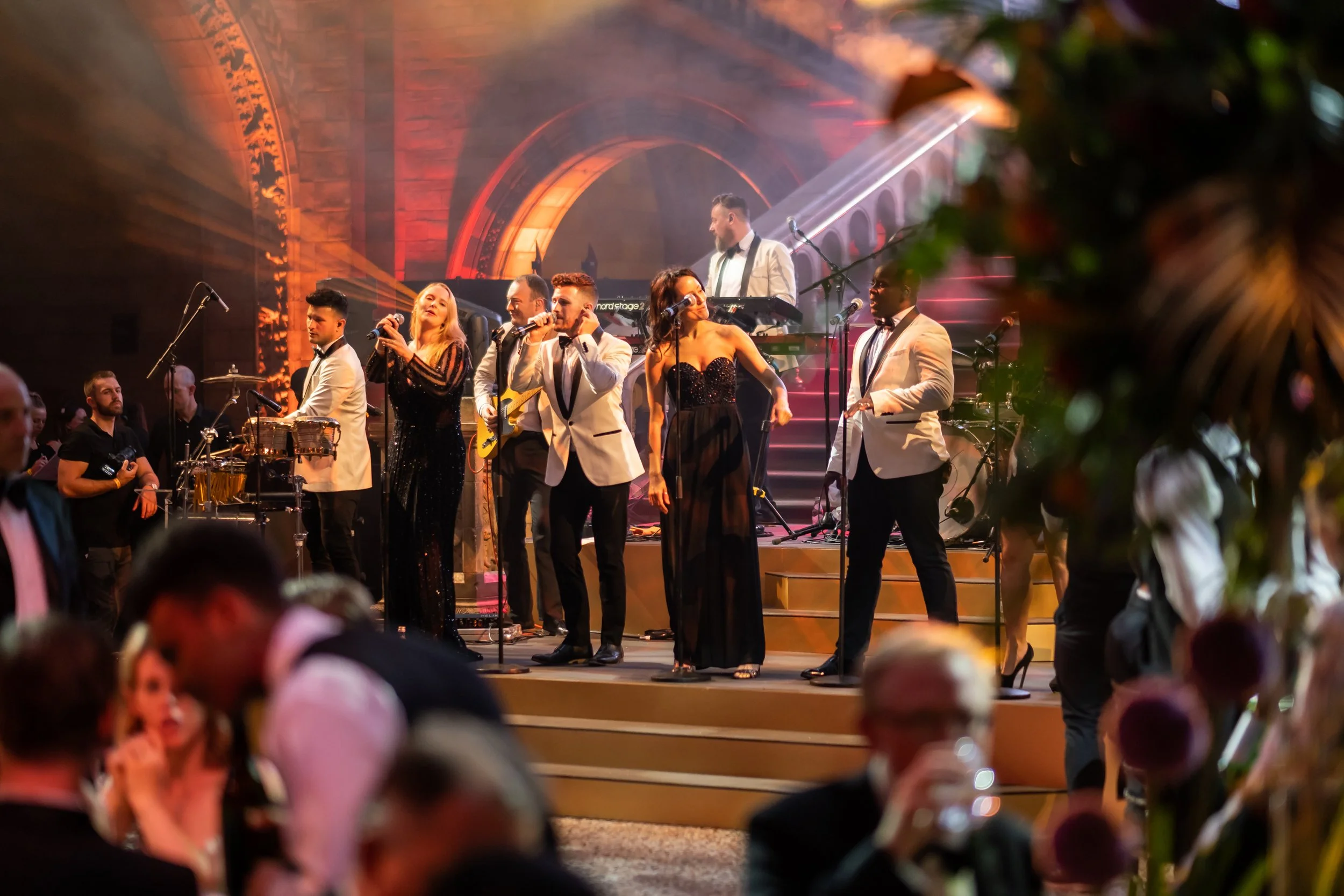 A live musical performance on stage with six singers dressed in formal attire, gold, black, and white, singing into microphones. The stage has dramatic lighting and an arched backdrop, with a keyboard and drums visible at The Natural History Museum