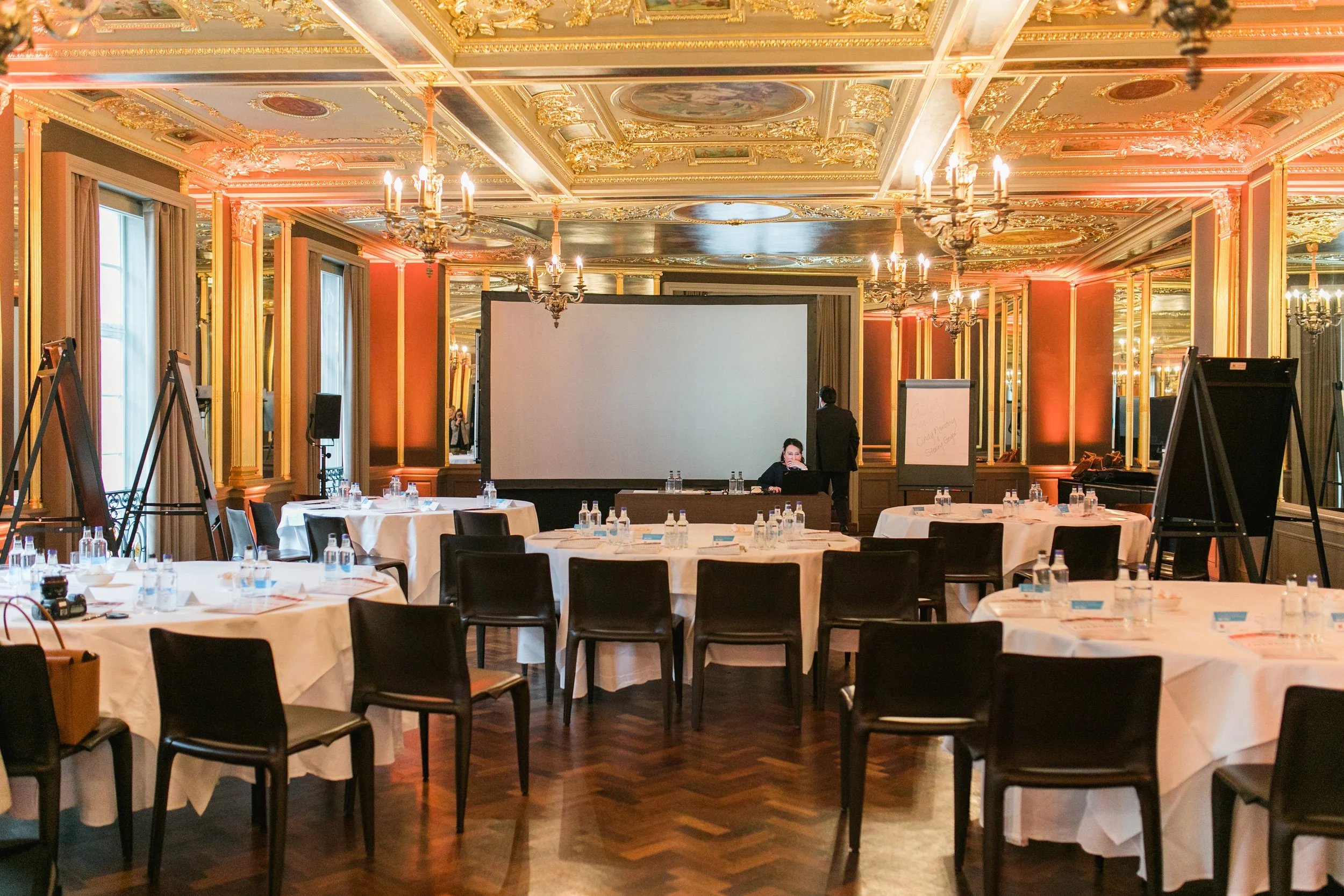 Elegant conference room with ornate gold-accented ceiling, chandeliers, large mirrors, and round tables set for a meeting or event.