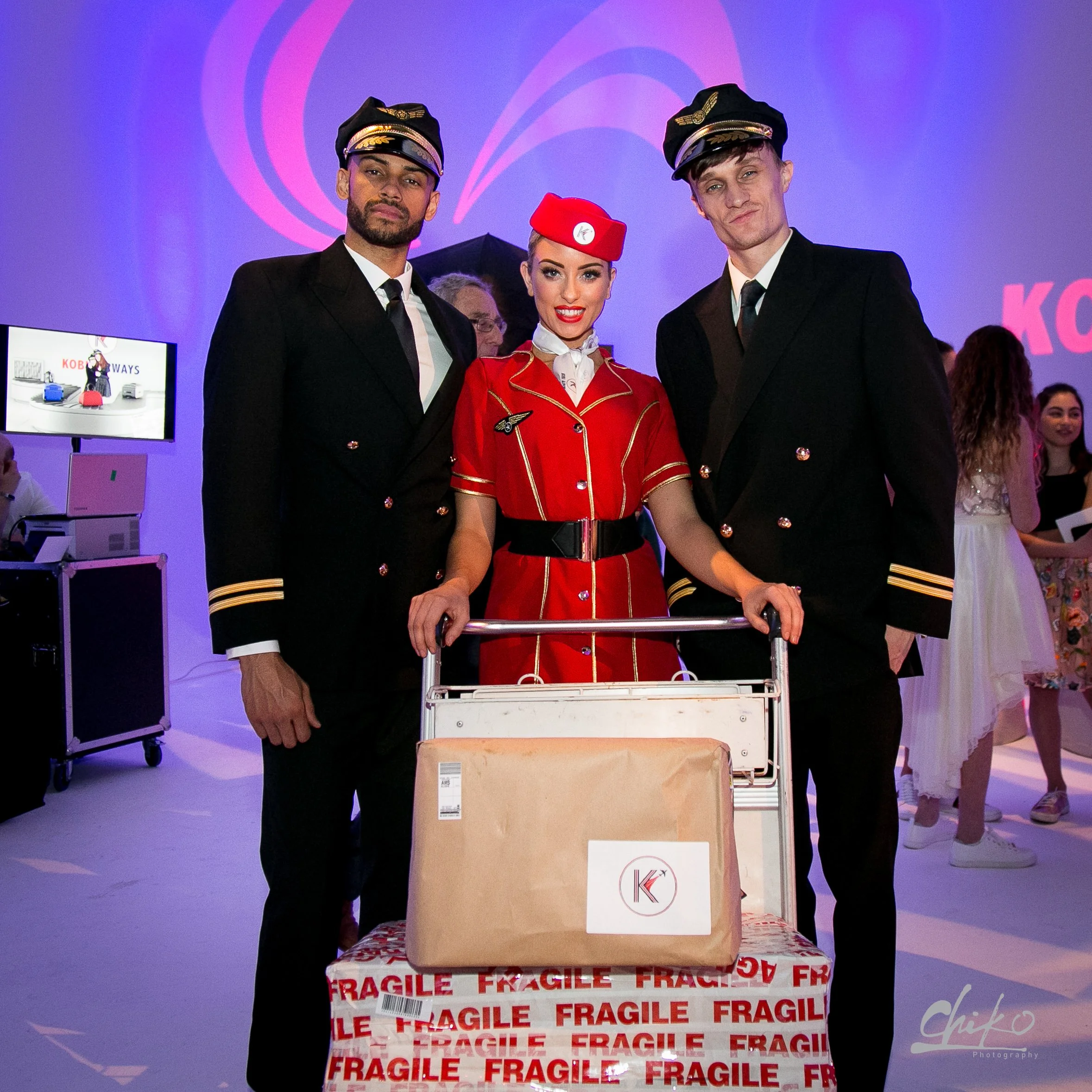 Three airline crew members dressed in uniform, standing behind a luggage trolley with a large, labeled box marked 'fragile' and a smaller box on top, at an airport or airport event.