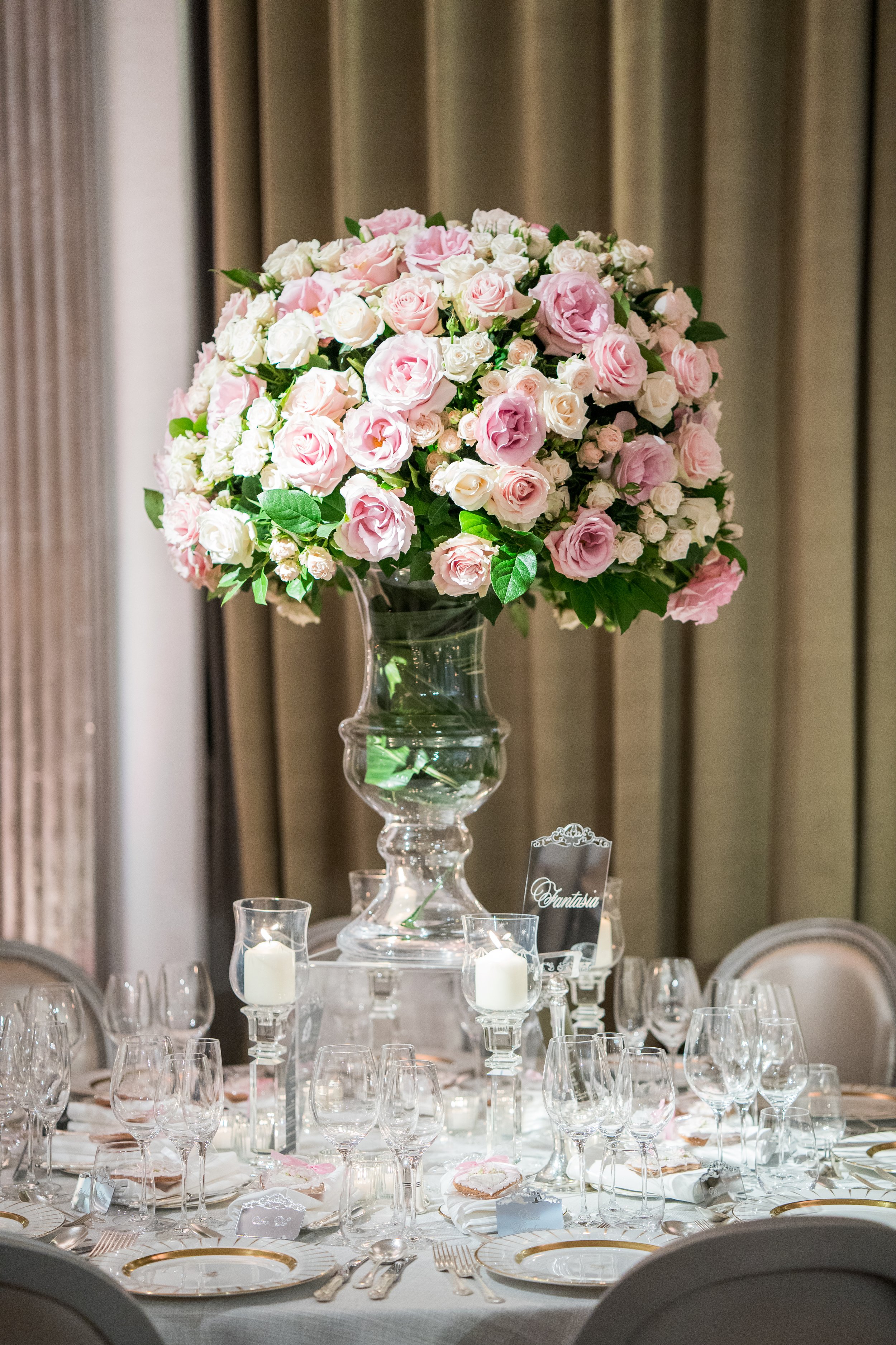Elegant dining table set with multiple wine glasses, plates with gold trim, and silverware, featuring a tall floral centerpiece of pink and white roses in a glass vase, under soft lighting in a formal setting.