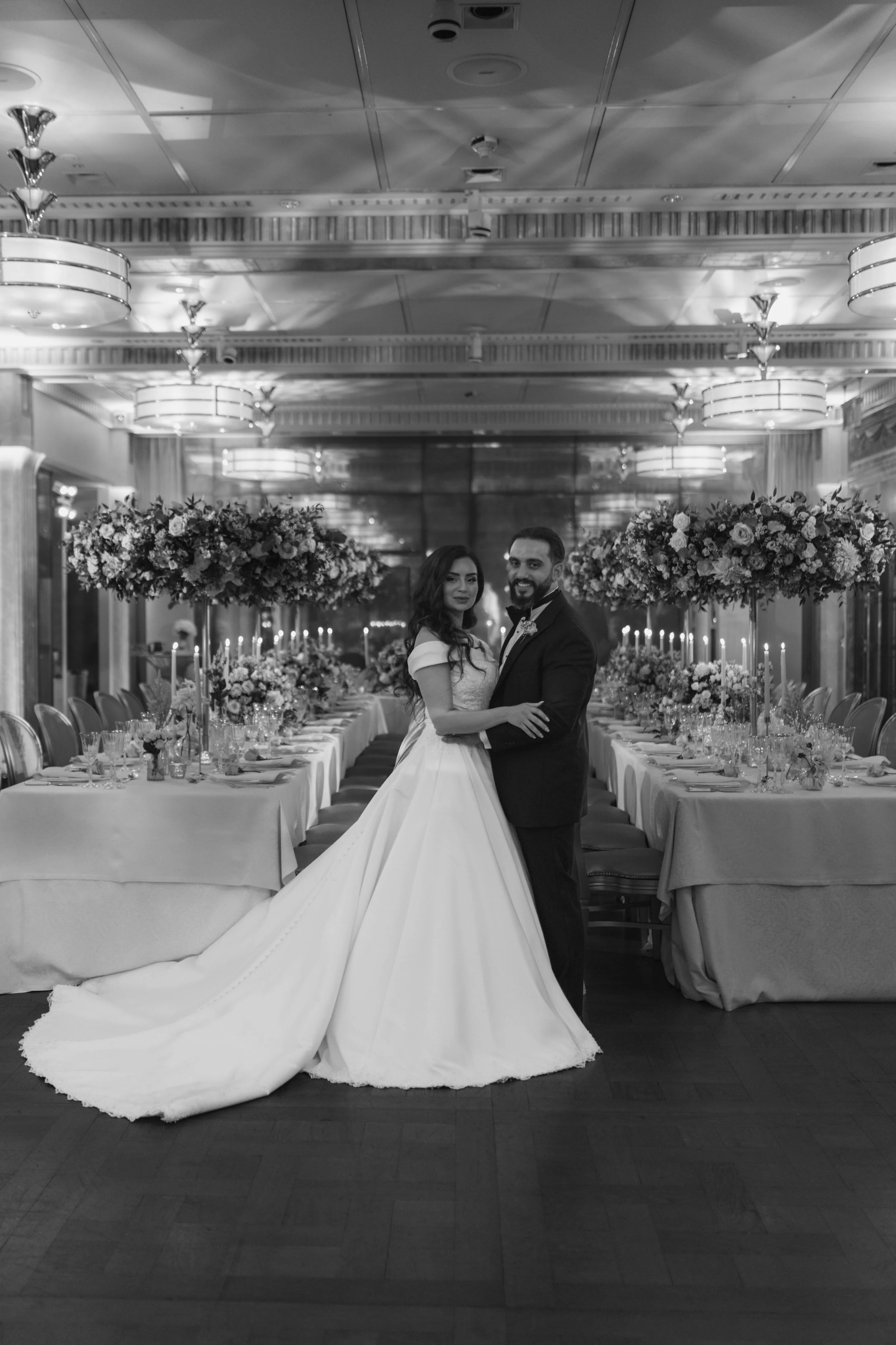 A bride and groom pose together in a banquet hall decorated with large flower arrangements and candles on a dine-in table. The bride wears a long white gown with a train, and the groom wears a tuxedo. The ceiling has decorative lighting fixtures.