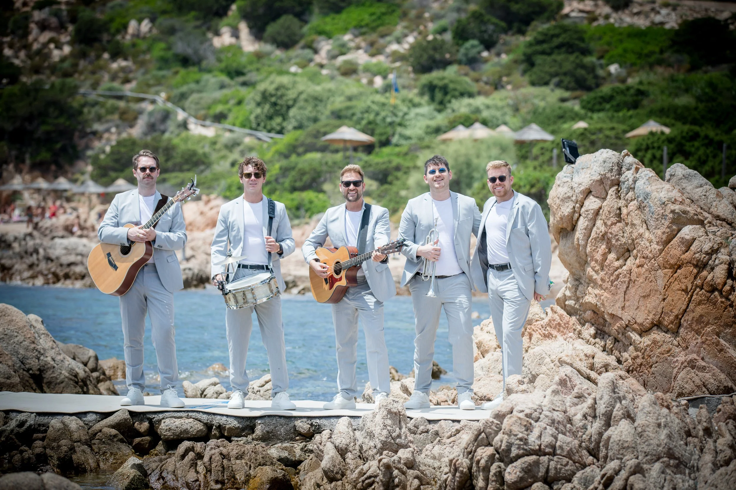 A group of five men dressed in white suits standing on a rocky shoreline with the ocean in the background. Some are holding musical instruments like guitars and a drum, and one has a trumpet. The scene is sunny with green hills and umbrellas in the d