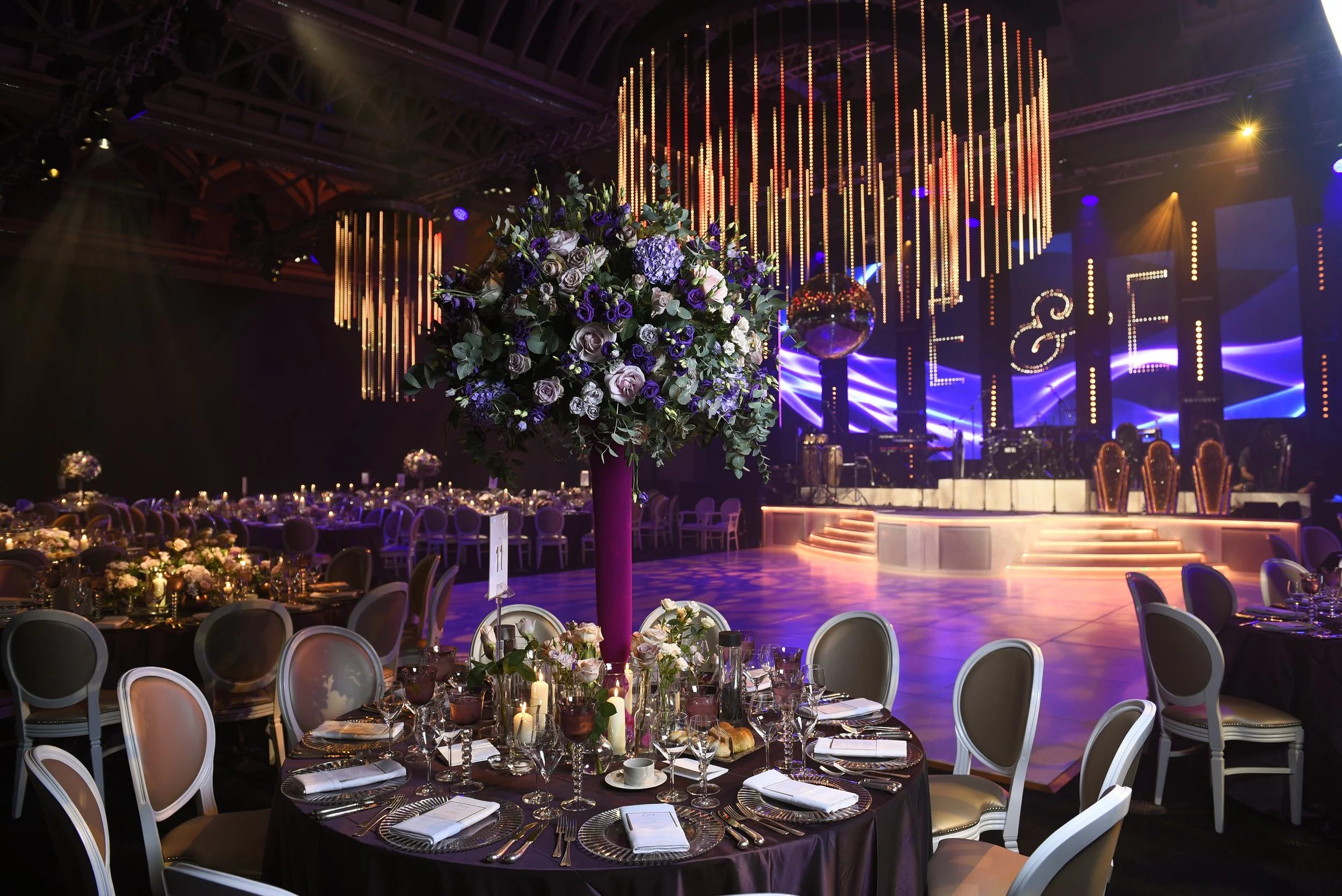 Elegant banquet hall decorated for a celebration with round tables, each set with fine tableware, white chairs, and floral centerpieces featuring roses and greenery. The background includes a stage with stage lights, a large disco ball, and a backdro