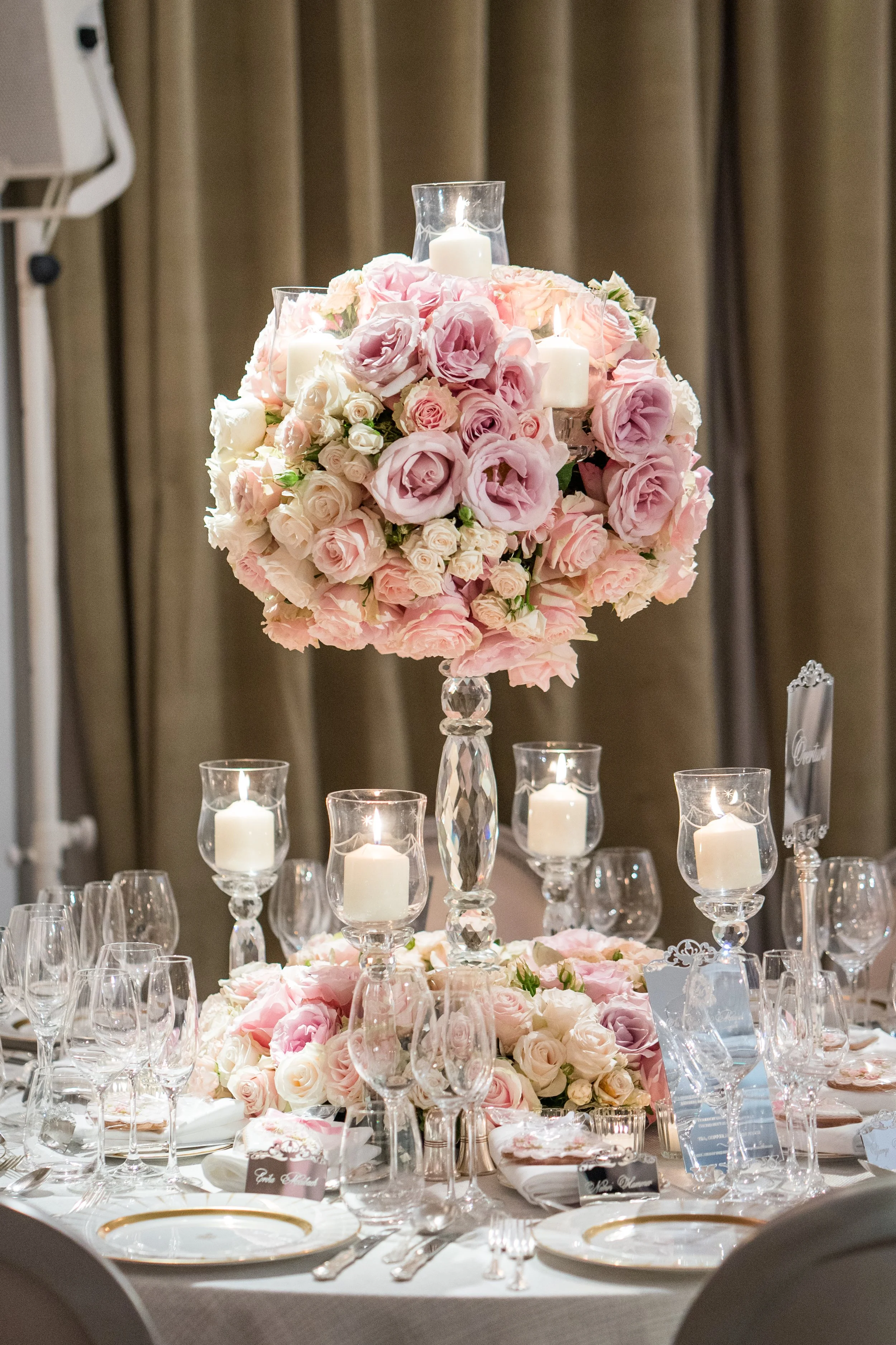 A wedding reception table with a tall floral centerpiece made of pink and white roses and hydrangeas, surrounded by candle holders with white candles, glassware, plates, and name cards on a white tablecloth.