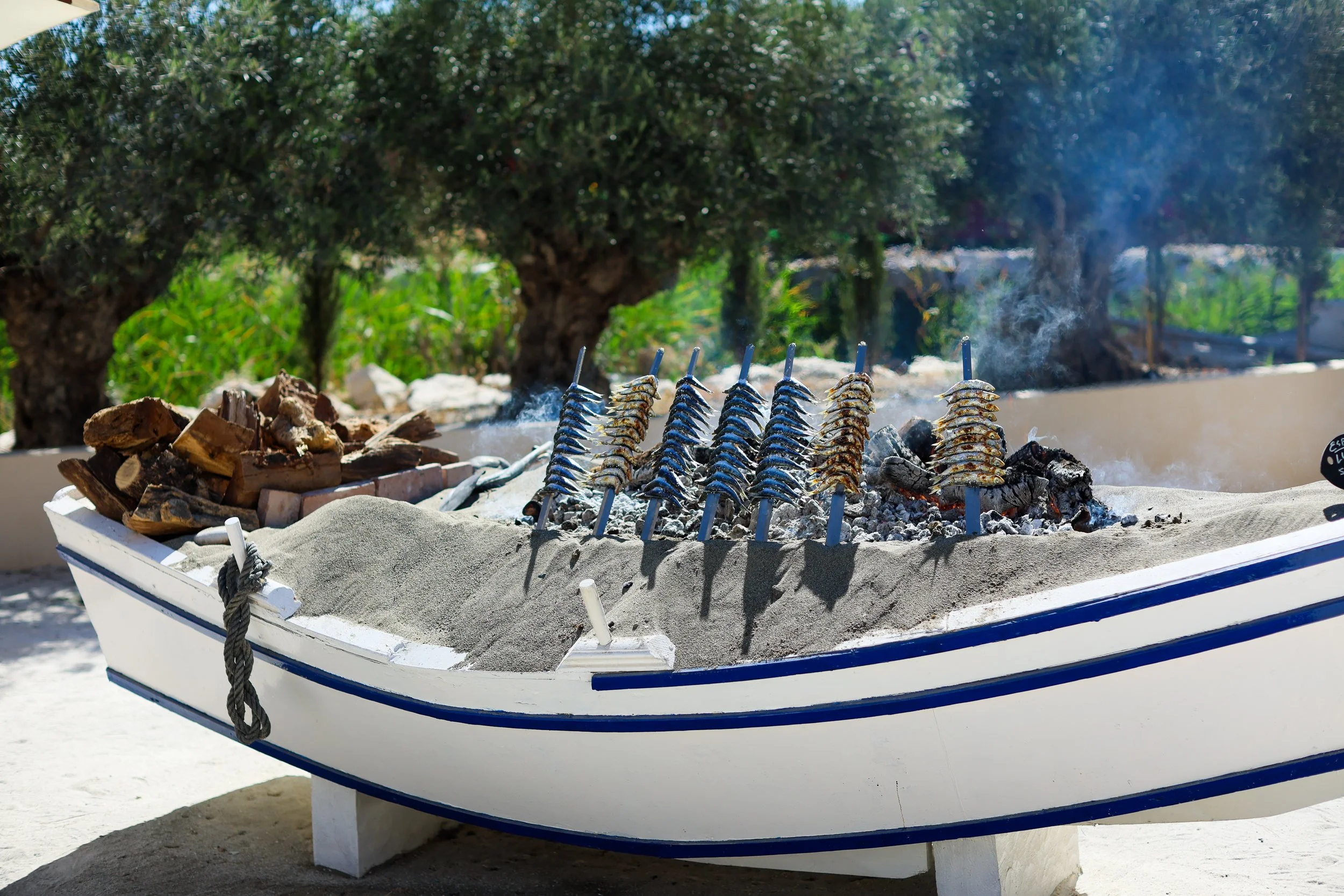 Grilled fish skewers cooking over a fire on a boat-shaped food stand, with a tree and greenery in the background.