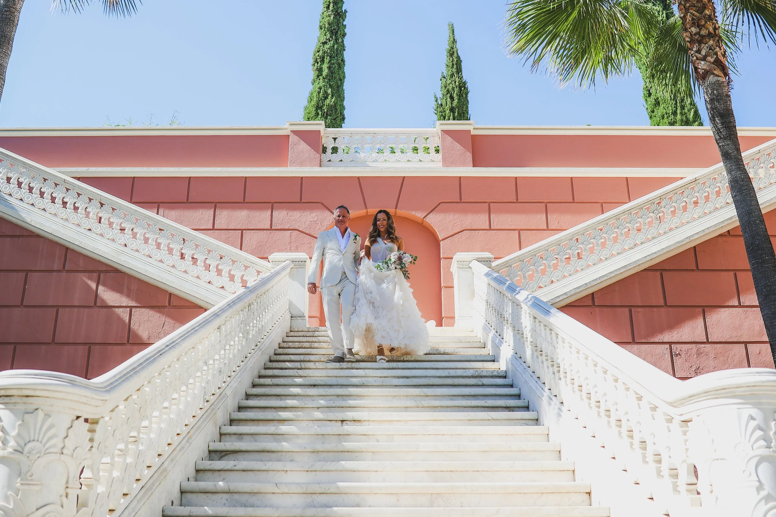 A bride and groom walking down a white staircase outside a pink building at Anantara Villa Padierna Resort in Marbella with palm trees and tall cypress trees in the background, on a clear sunny day.