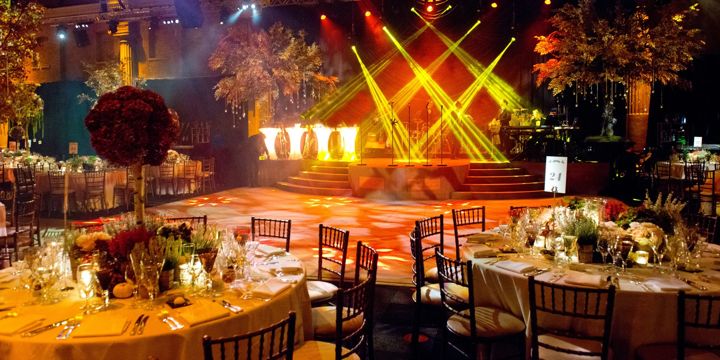 Indoor event space decorated with round tables covered in white tablecloths, set with glassware and floral centerpieces, with a stage illuminated by yellow and red lights, and surrounded by trees and chairs.