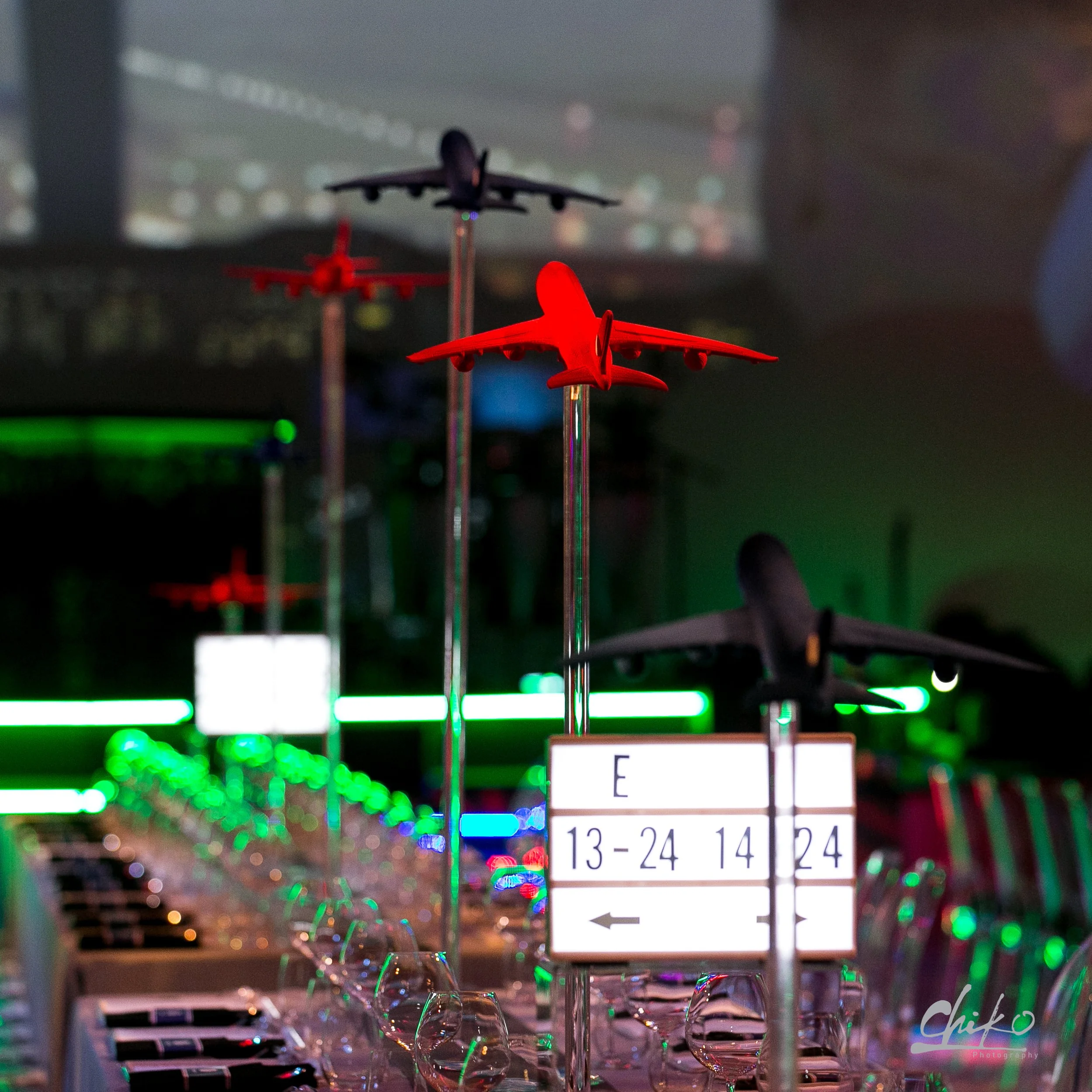 Table setting with wine glasses, a sign indicating table numbers, and a blurred cityscape view of a plane in flight seen through a window.
