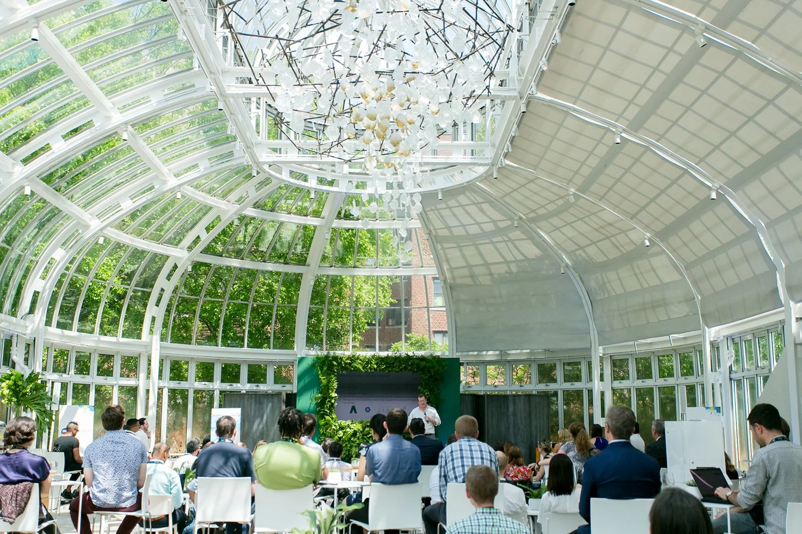 A conference or presentation taking place inside a bright, glass-roofed conservatory-style building filled with natural light. A speaker is standing at the front, addressing an audience seated and standing, with large green plants and a decorative ch