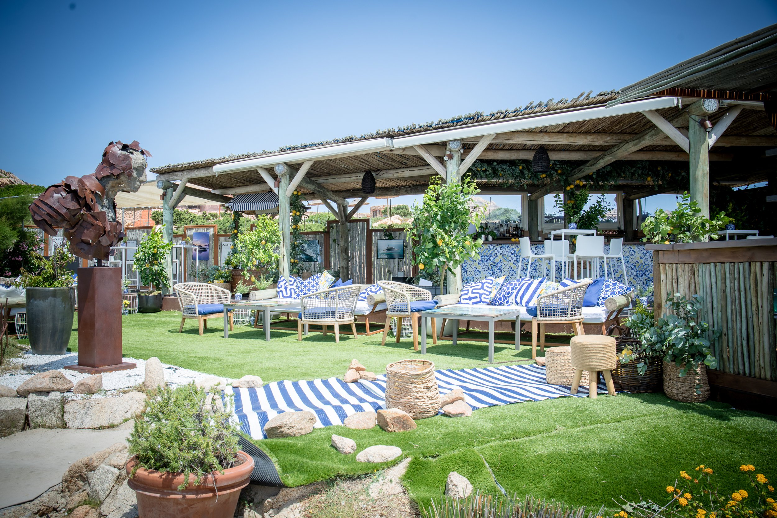 An outdoor patio with a covered seating area, surrounded by plants and garden decor, including a bronze bust sculpture and framed pictures.