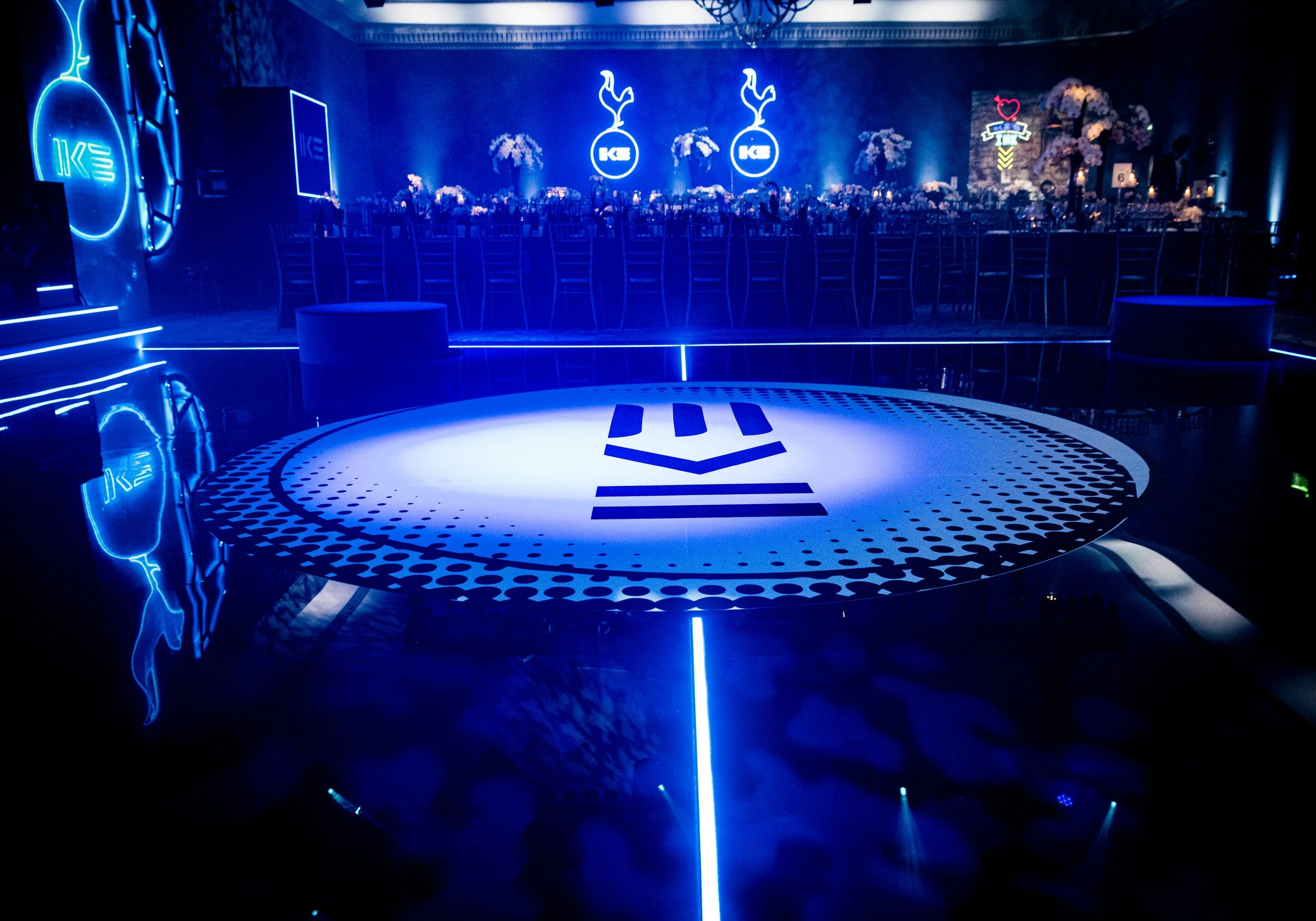 A nightclub scene with blue neon lighting, a circular dance floor, and stage area with chairs, flowers, and neon decorations.