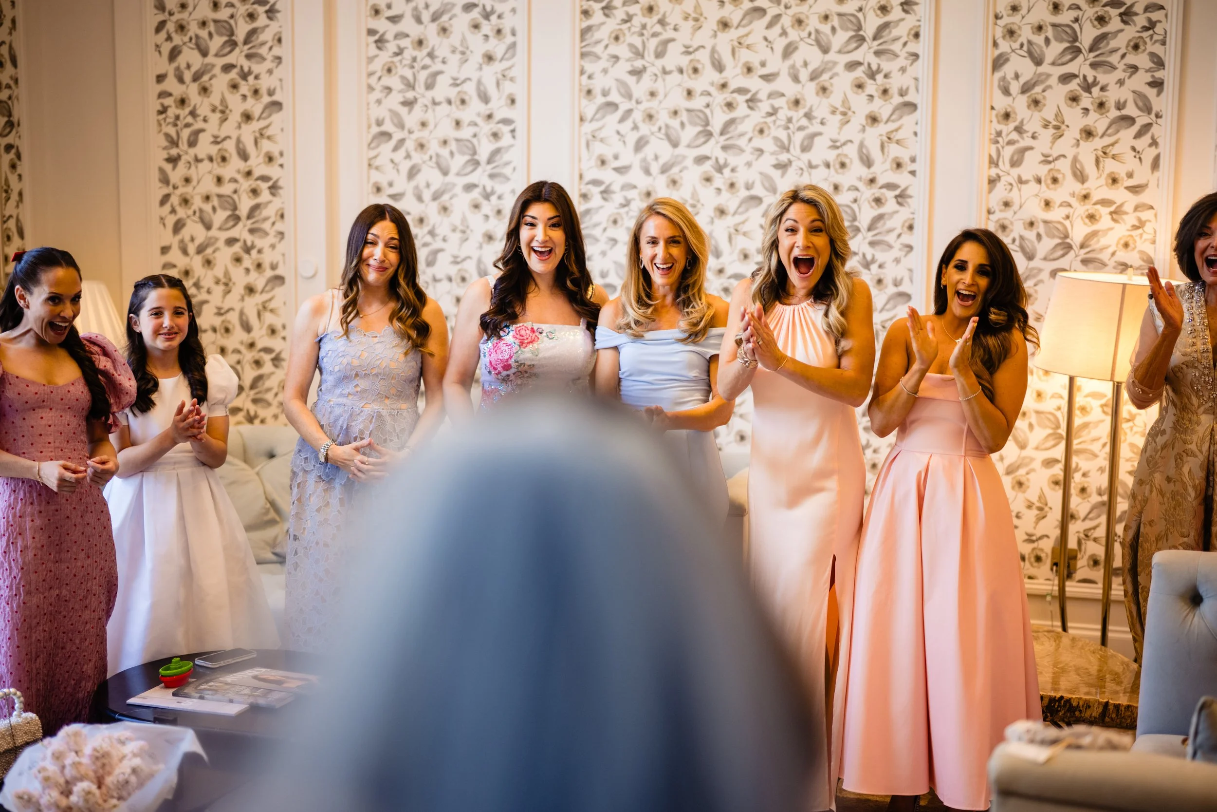 Group of women and girls dressed in elegant dresses, smiling and celebrating indoors with a floral wallpaper background. Fairmont Windsor Park