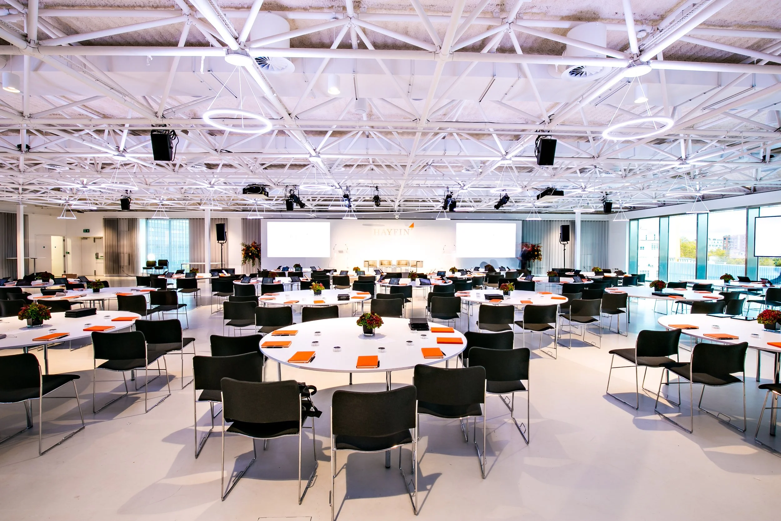 A large conference room or event space with round tables arranged for a meeting or seminar. Each table has orange folders, notepads, and pens. There are black chairs around the tables. The room has large windows on one side, allowing natural light, a