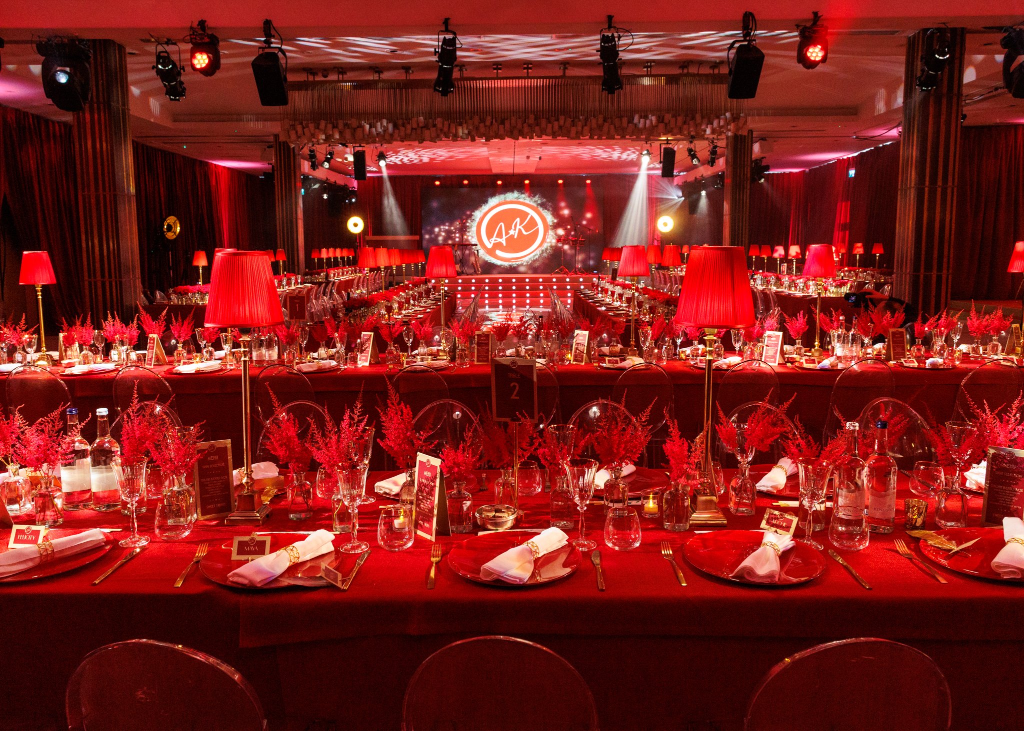 A large banquet hall decorated for an event, with tables set with white napkins, gold utensils, glasses, and red floral centerpieces. The room features red curtains, red lampshades, and a stage with lighting and a large circular logo with the initials 'A & K' in the background.