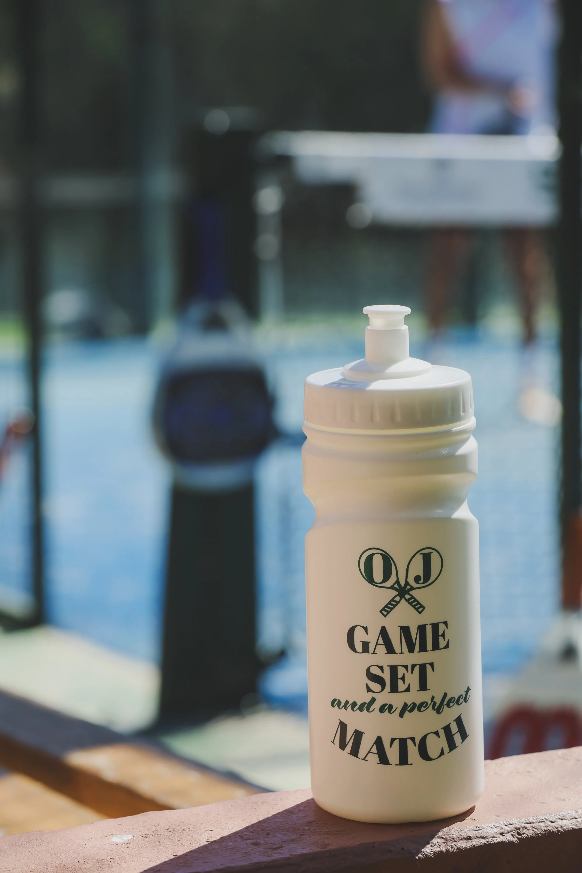 A white water bottle with black lettering reading 'Game Set and a Perfect Match' sits on a pink brick ledge outdoors, with blurred objects and a person in the background.