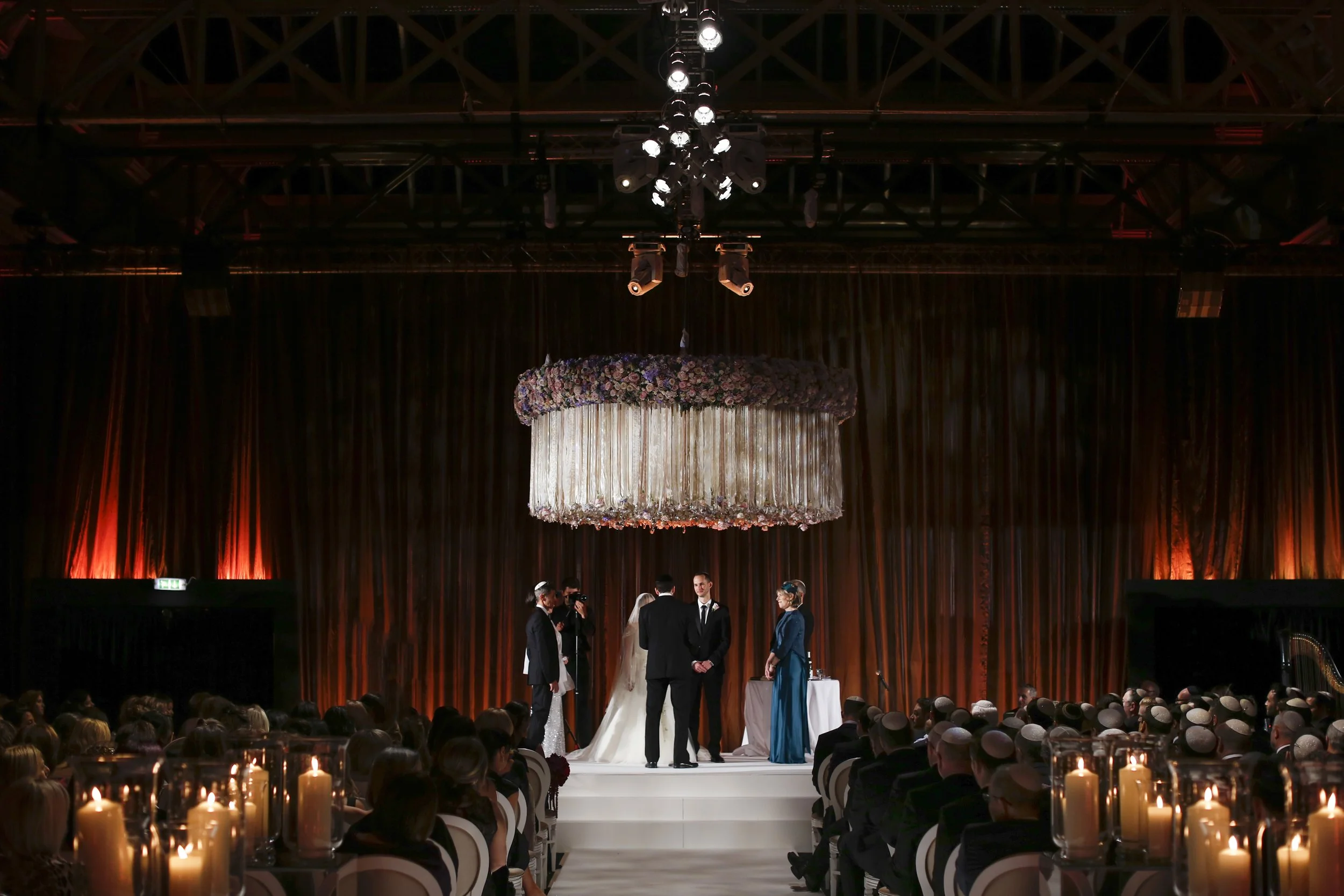 A wedding ceremony taking place indoors, with a bride and groom standing at the altar, surrounded by family and friends. A large floral arrangement hangs from the ceiling, and candles are lit along the aisle.